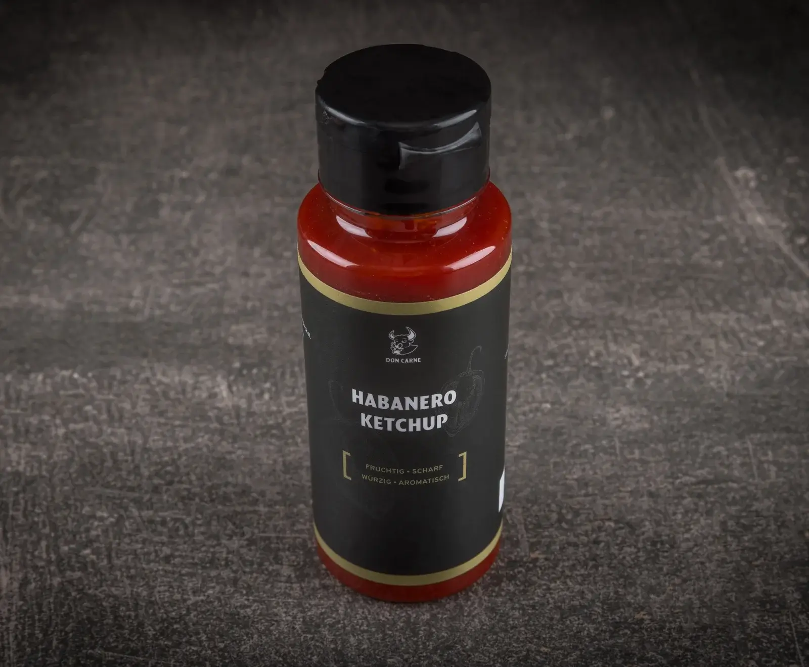 Eine rote Flasche Don Carne Habanero Ketchup mit schwarzem Etikett und goldenen Akzenten steht auf dunklem, strukturiertem Untergrund. Auf dem Etikett steht "HABANERO KETCHUP" und "FRUCHTIG • SCHARF WÜRZIG • AROMATISCH".