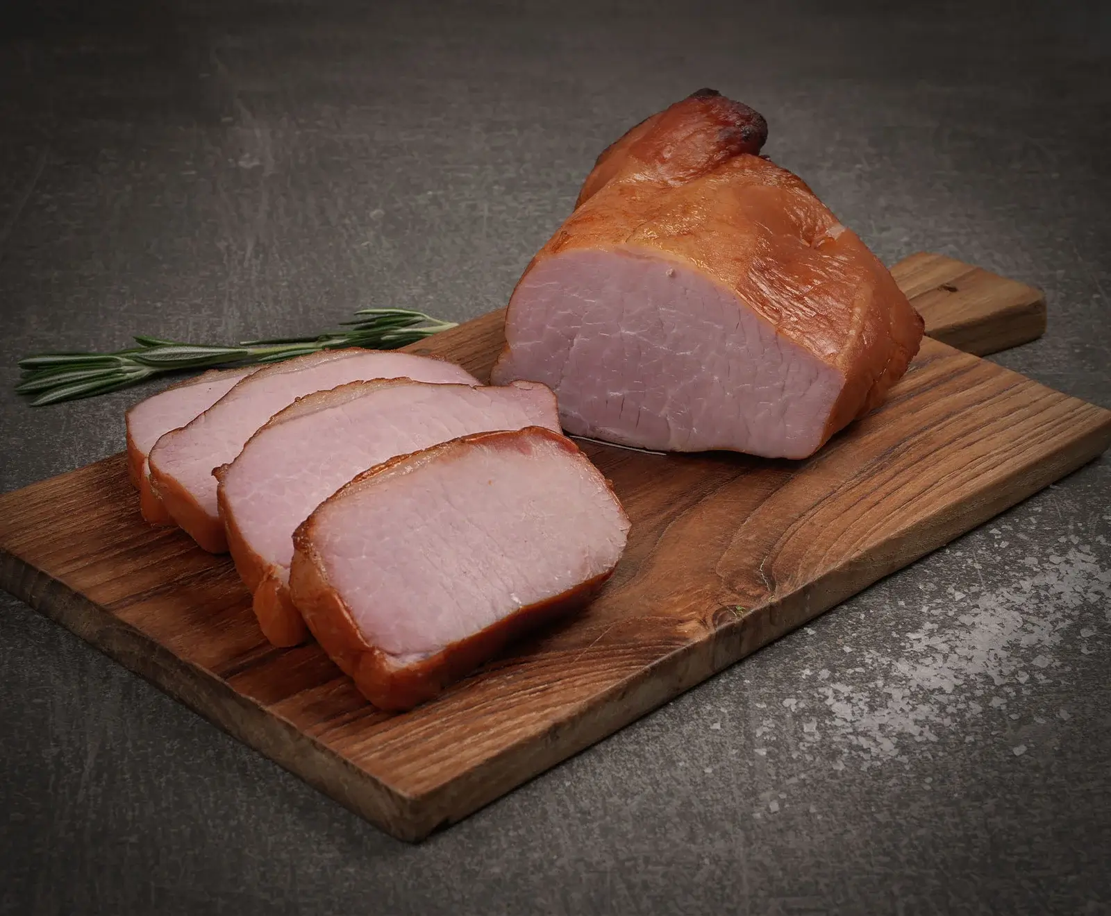 Ein Stück geräucherter Kassler-Lachsbraten, teilweise in Scheiben geschnitten, liegt auf einem Holzschneidebrett. Daneben ein Rosmarinzweig auf dunklem Untergrund mit Salz.