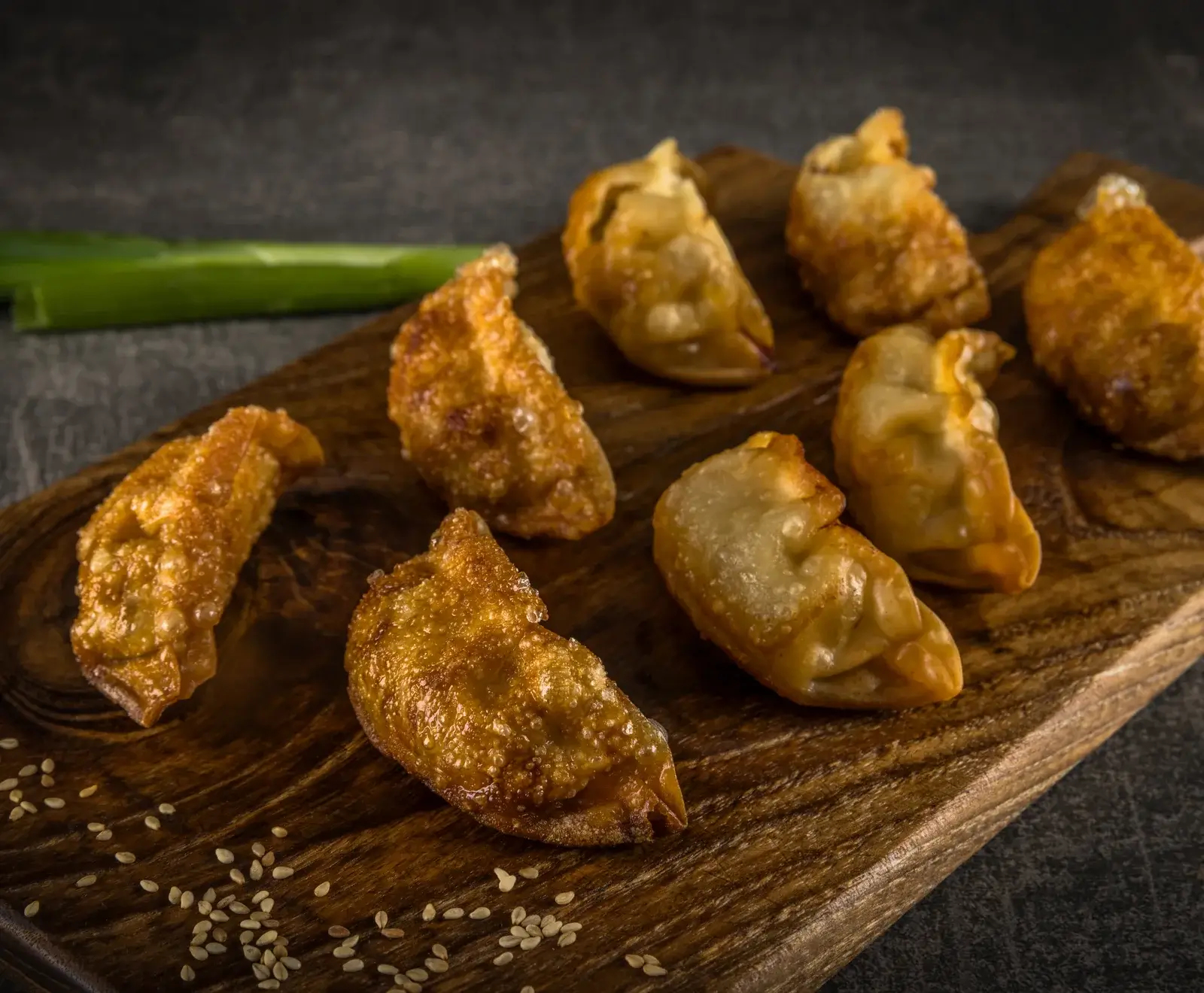 Mehrere goldbraun gebratene Teigtaschen (Gyoza) liegen auf einem dunklen Holzbrett. Einige Sesamsamen sind verstreut, im Hintergrund ist eine grüne Lauchzwiebel zu sehen.