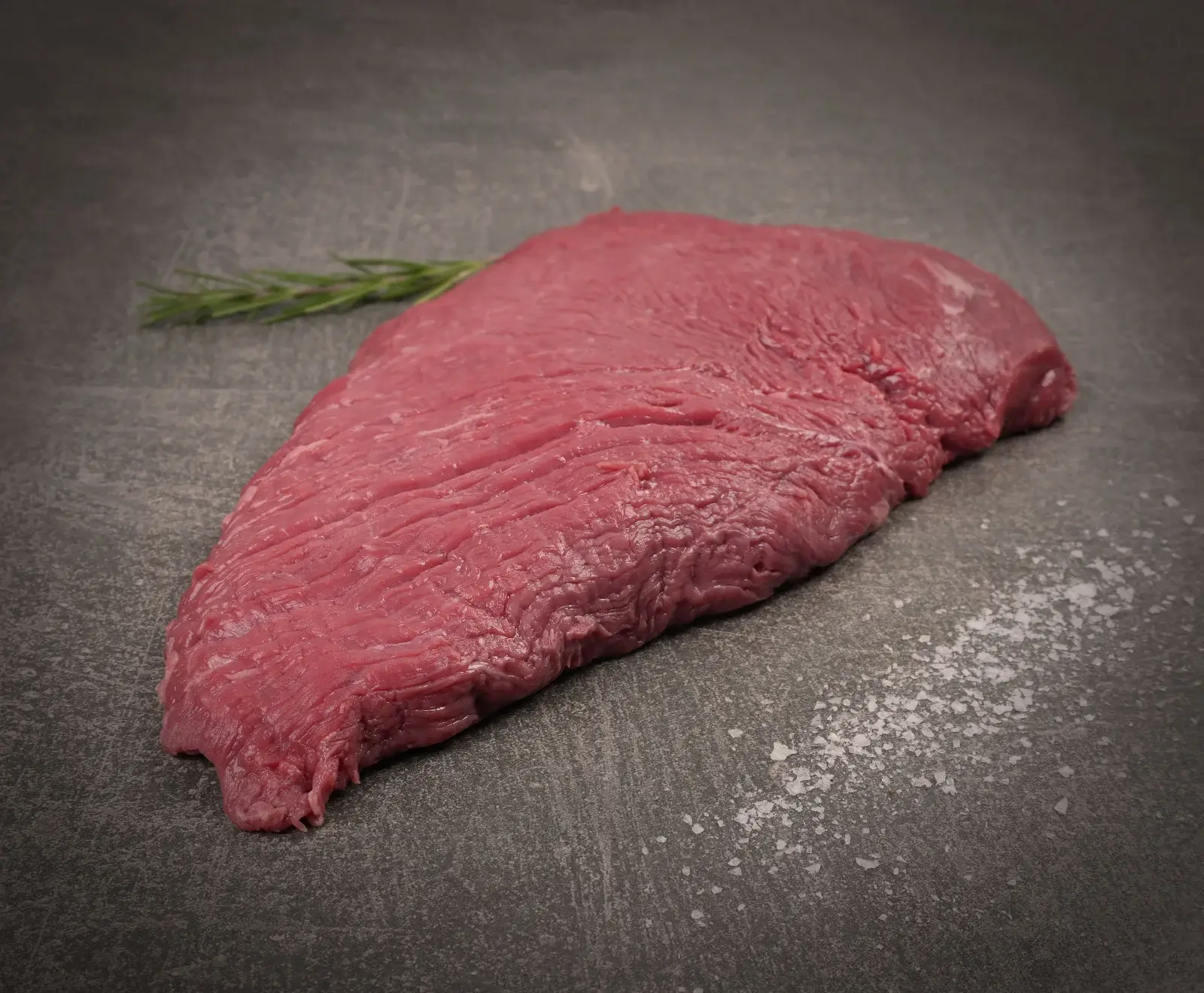 Ein rohes, mageres Stück Rindfleisch liegt auf einer dunklen, strukturierten Oberfläche. Im Hintergrund ist ein Rosmarinzweig zu sehen, im Vordergrund grobes Salz verstreut.