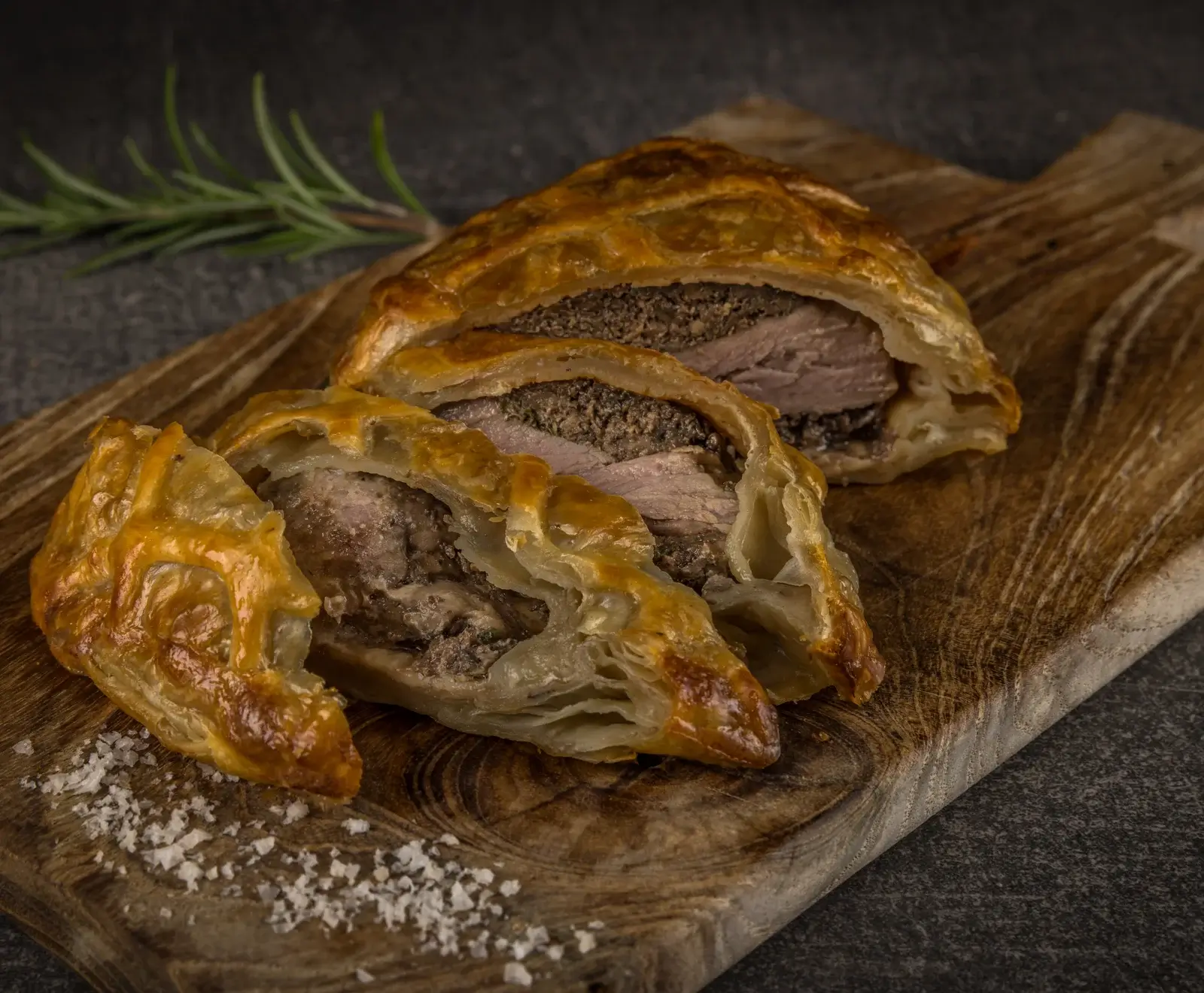 Ein aufgeschnittenes Beef Wellington auf einem rustikalen Holzbrett, das die Schichten aus goldbraunem Blätterteig, dunkler Pilzfarce und zartrosa Rinderfilet zeigt. Grobes Salz und Rosmarin sind sichtbar.