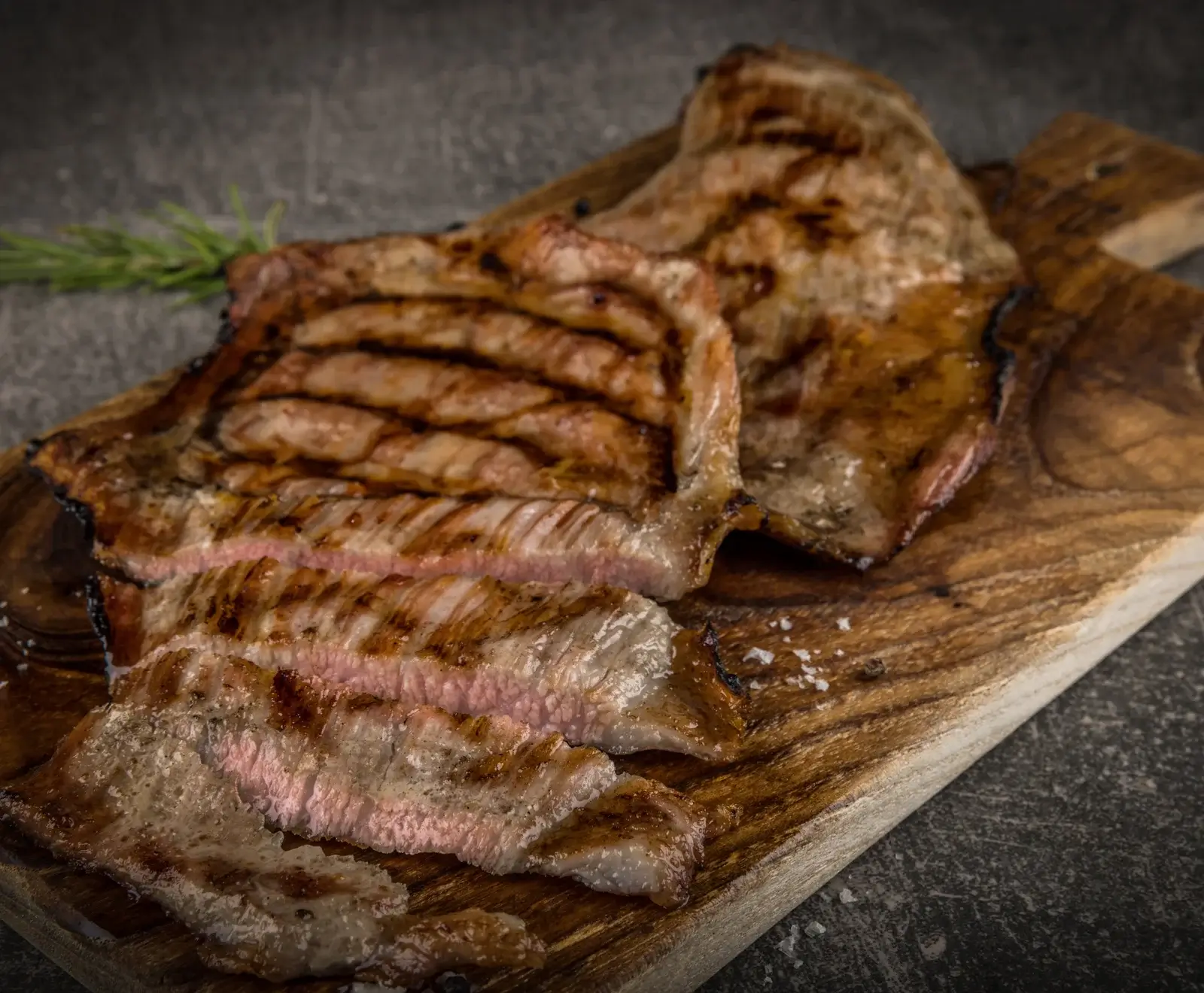 Ein saftiges, gegrilltes Steak, teilweise in Scheiben geschnitten, mit rosa Kern, deutlichen Grillspuren und Meersalz auf einem Holzbrett. Rosmarin im Hintergrund.