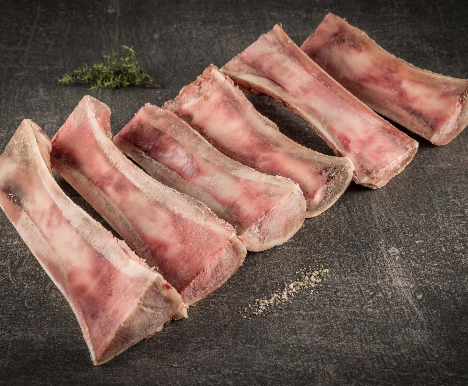 Sechs gefrorene Markknochen liegen diagonal auf einer dunklen Steinplatte. Ein Zweig Thymian und eine Prise grobes Salz mit Pfeffer sind daneben zu sehen.