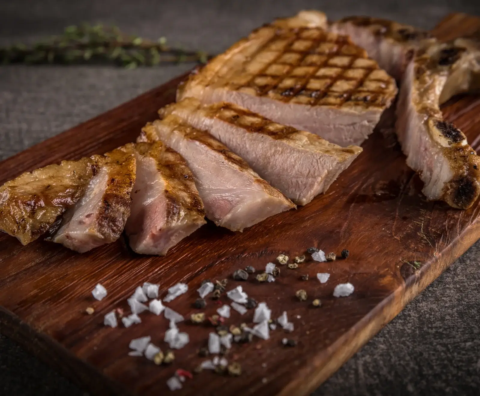 Ein gegrilltes Schweinekotelett, teilweise in Scheiben geschnitten, liegt auf einem dunklen Holzbrett. Grobes Salz und bunte Pfefferkörner sind darauf verstreut. Im Hintergrund ist ein Kräuterzweig zu sehen.