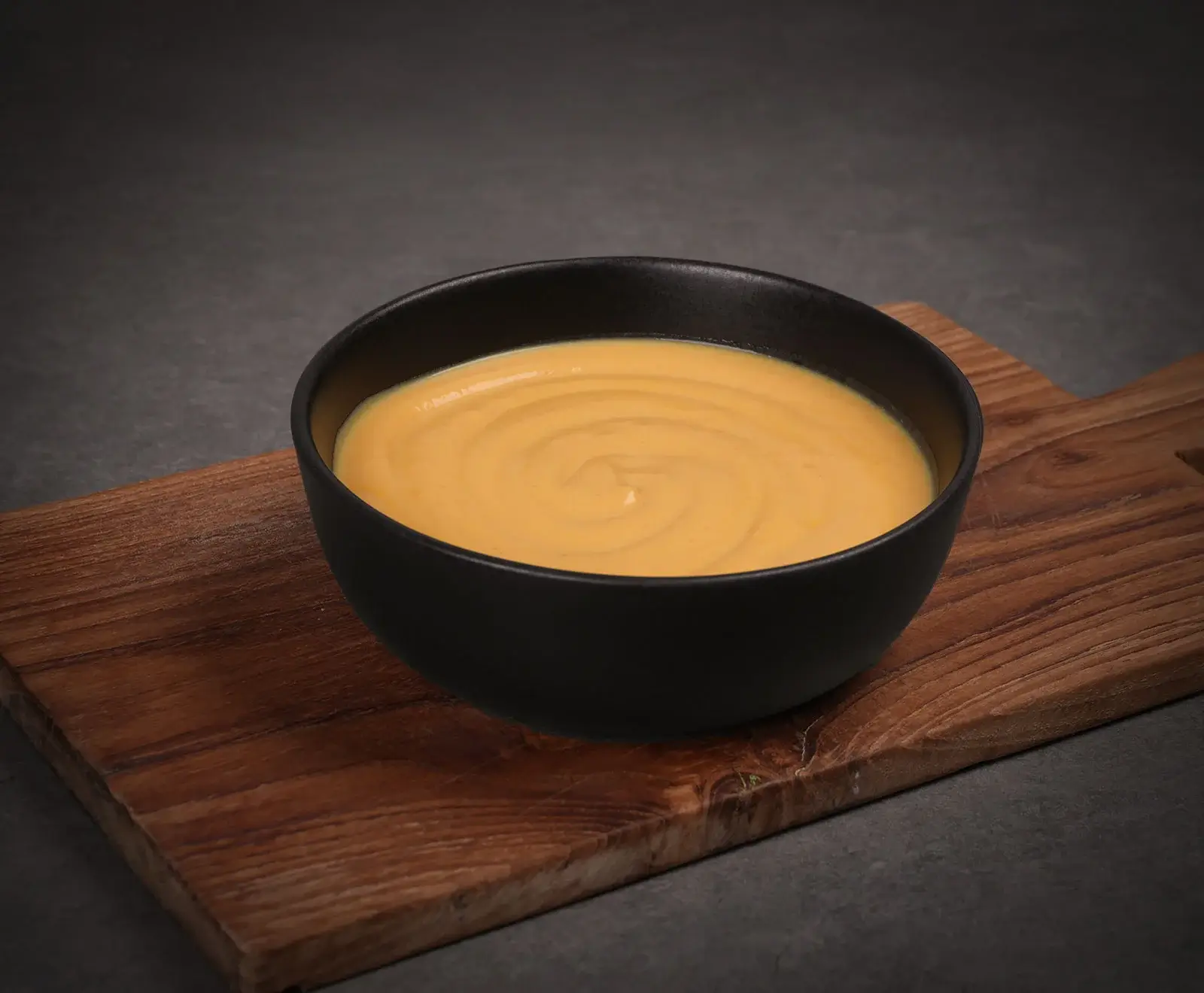 Eine schwarze Schale mit einer glatten, goldgelben, cremigen Suppe oder Soße, die auf einem rustikalen Holzbrett steht. Der Hintergrund ist dunkelgrau.