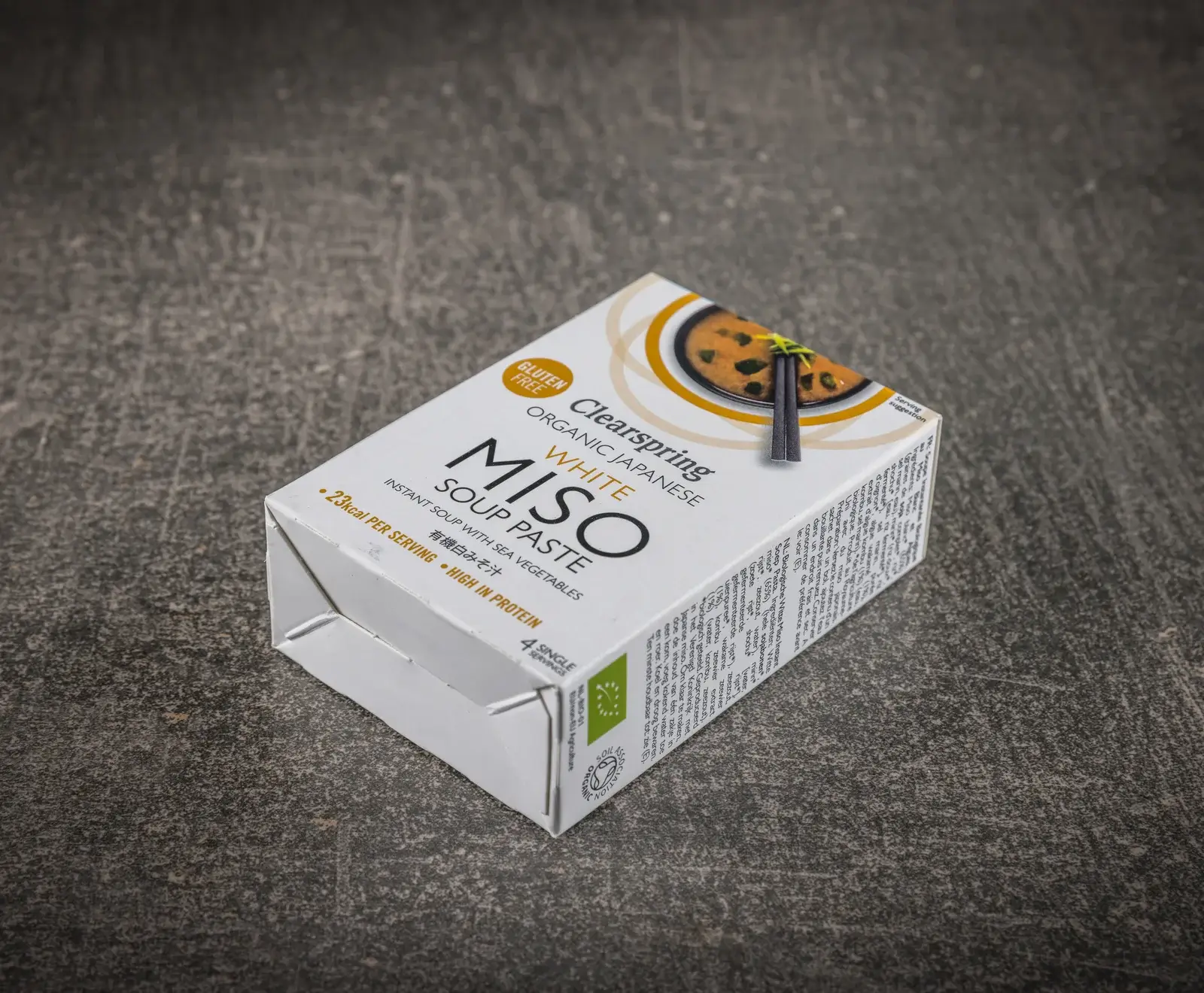 Eine weiße Packung Clearspring Bio Japanische Weiße Miso Suppenpaste, glutenfrei, mit Meeresgemüse, 23kcal pro Portion, proteinreich, für 4 Portionen, auf dunklem Untergrund. Oben ist ein Bild von Misosuppe mit Essstäbchen.