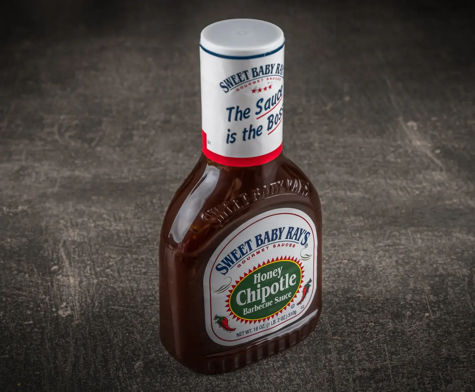 Eine dunkelbraune Flasche Sweet Baby Ray's Honey Chipotle Barbecue Sauce mit weißem Etikett und grüner Aufschrift "Honey Chipotle" auf dunklem, strukturiertem Hintergrund.