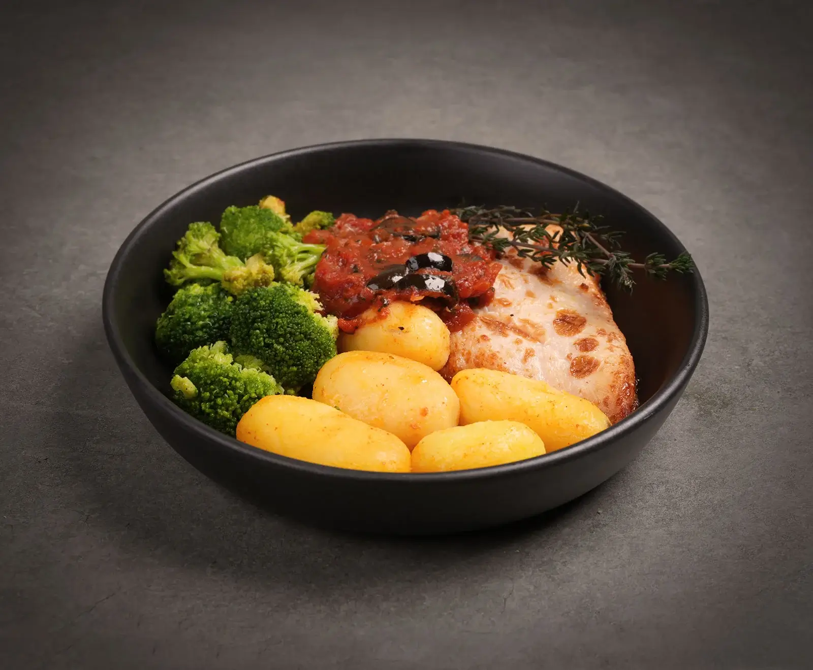 Eine schwarze Schale mit gebratenem Fleisch, kleinen Kartoffeln, Brokkoli und Tomatensauce mit Oliven, garniert mit Thymian, auf dunklem Hintergrund.
