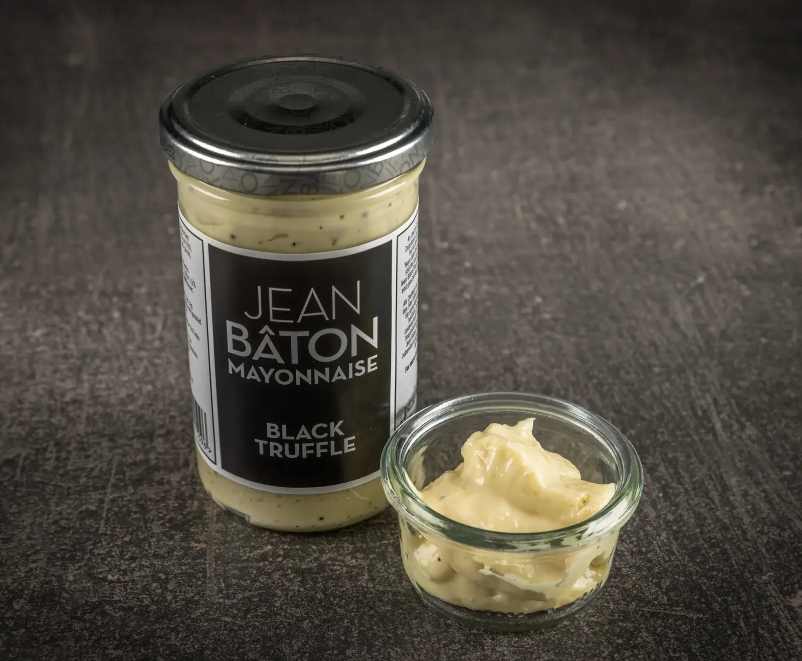 Ein Glas Jean Bâton Mayonnaise mit schwarzem Trüffel steht neben einem kleinen Glasgefäß, das die cremige, hellgelbe Mayonnaise mit dunklen Trüffelstücken enthält, auf dunklem Untergrund.