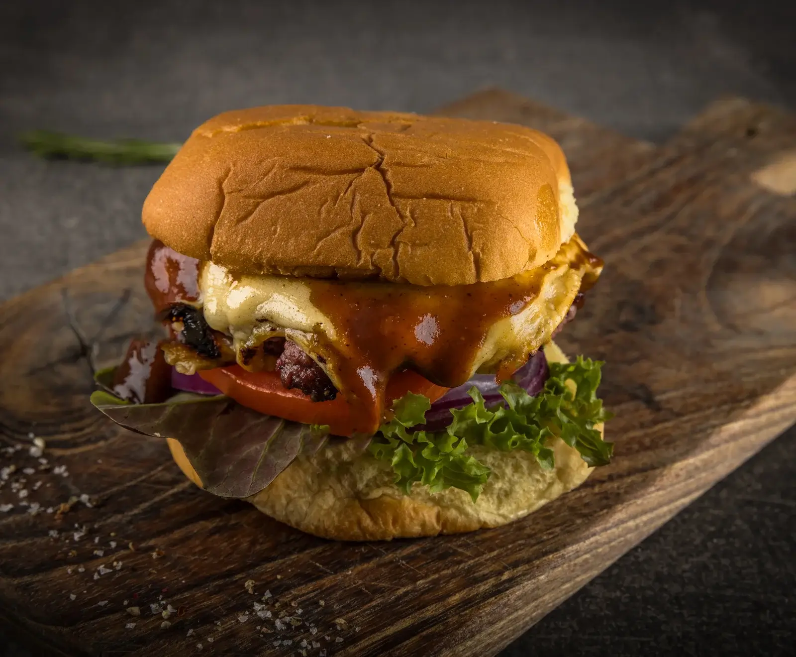 Ein appetitlicher Burger mit geschmolzenem Käse, reichlich brauner Sauce, Salat, Tomate und roten Zwiebeln auf einem rustikalen Holzbrett, mit Salz bestreut.