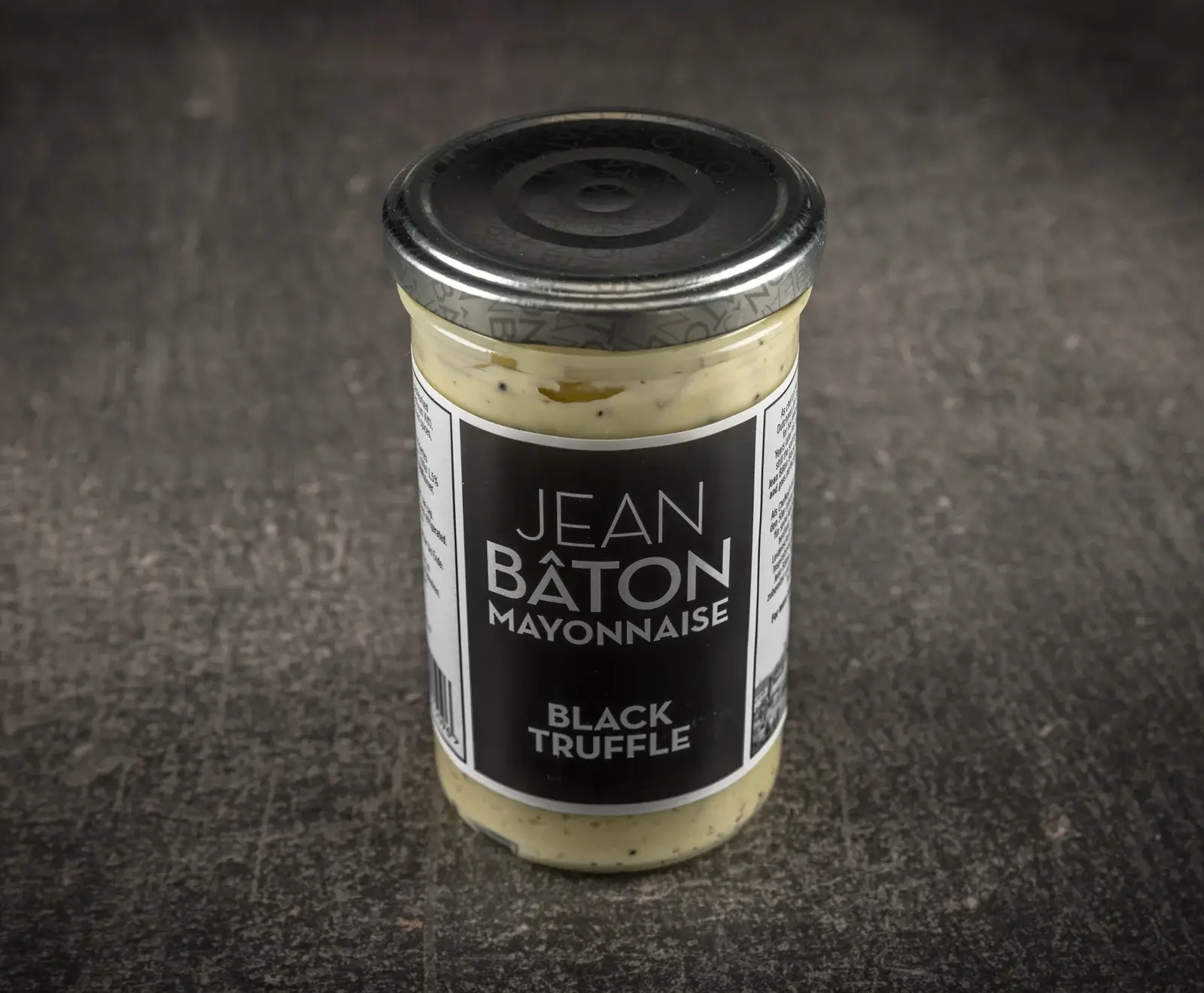 Ein Glas Jean Bâton Mayonnaise mit schwarzem Trüffel. Die helle Mayonnaise mit dunklen Sprenkeln hat einen silbernen Deckel und steht auf einem dunklen, strukturierten Untergrund.