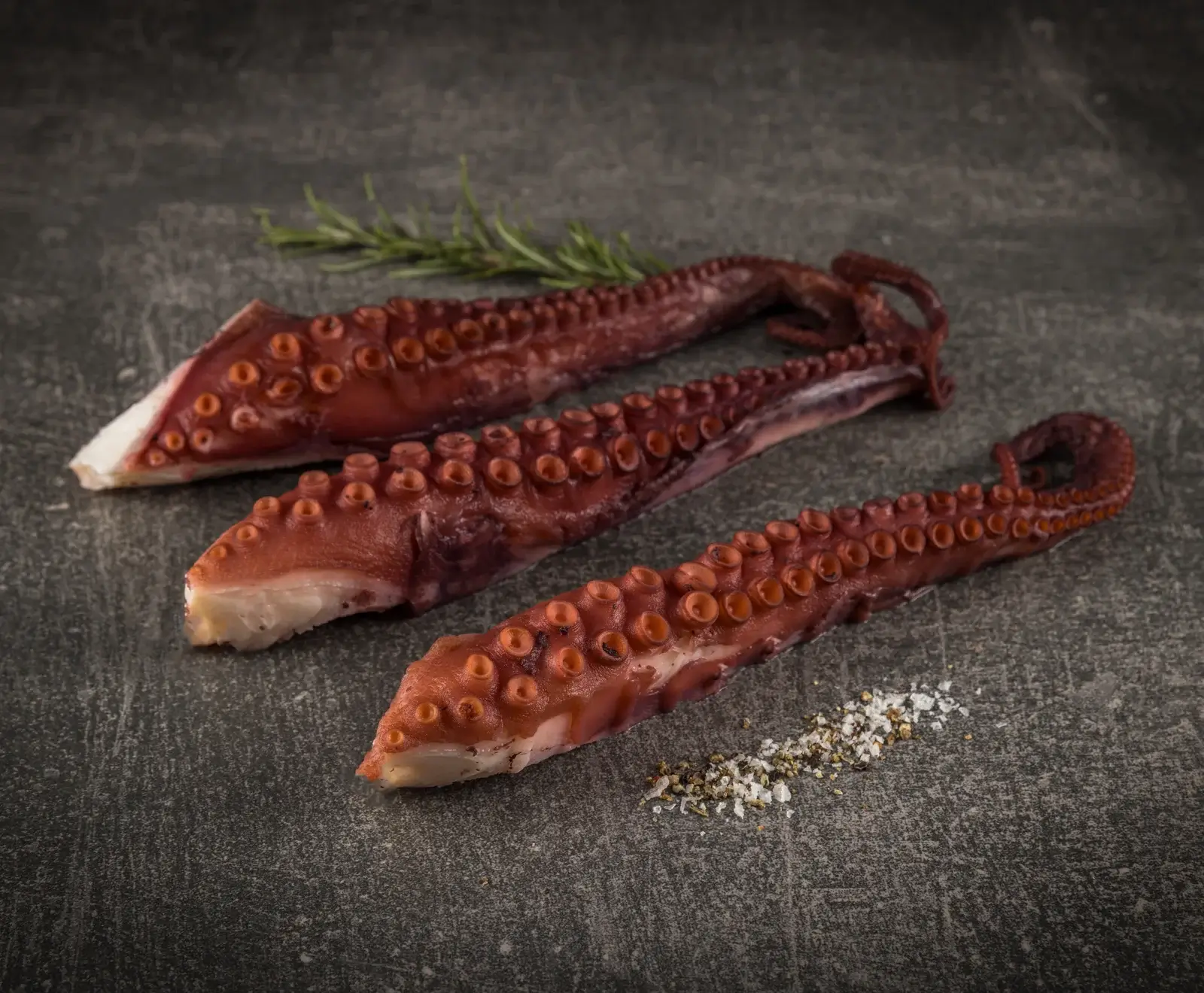 Drei gekochte, rötlich-braune Oktopusarme mit Saugnäpfen liegen auf einer dunklen Steinplatte. Im Hintergrund ist ein Rosmarinzweig, im Vordergrund eine Prise Salz und Pfeffer.
