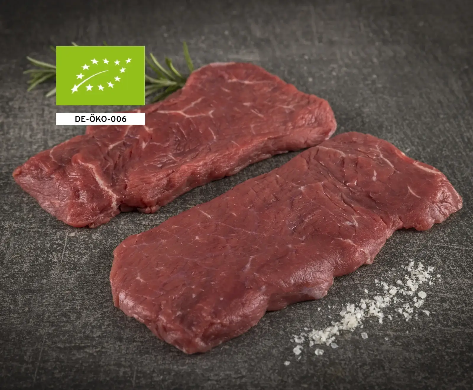Drei rohe Bio-Rindersteaks auf dunklem Hintergrund mit Salz, Rosmarin und EU-Bio-Logo