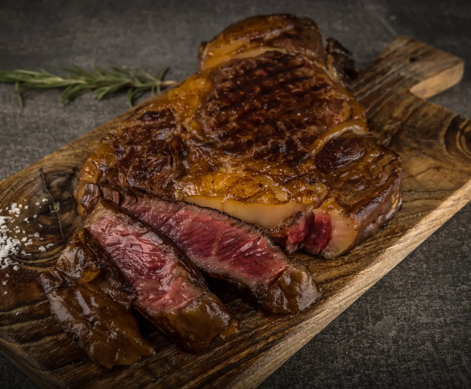 Gegrilltes Ribeye-Steak mit perfekter Kruste, in Scheiben geschnitten und auf Holzbrett serviert, mit Rosmarin garniert.