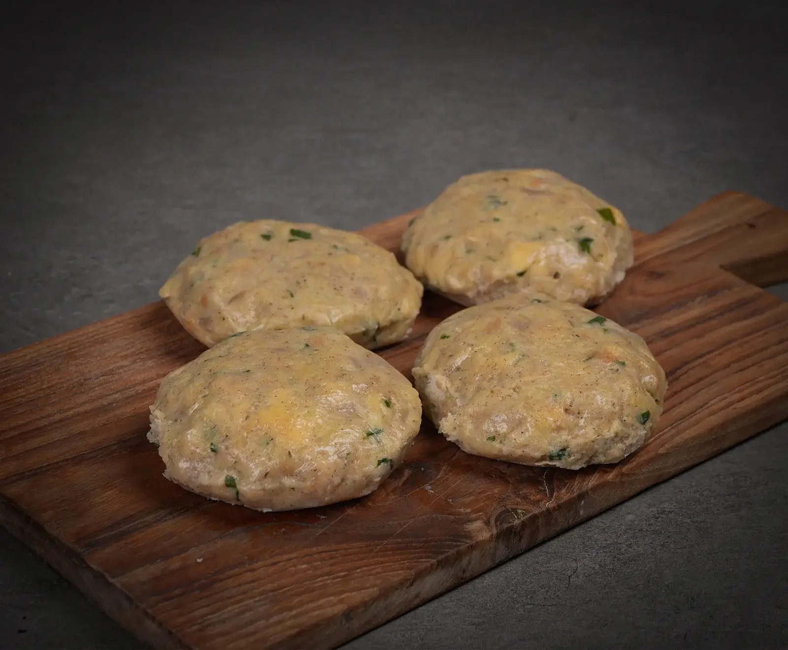 Vier ungegarte, runde Knödel mit sichtbaren Kräutern und kleinen gelben Stücken liegen auf einem dunklen Holzschneidebrett vor grauem Hintergrund.