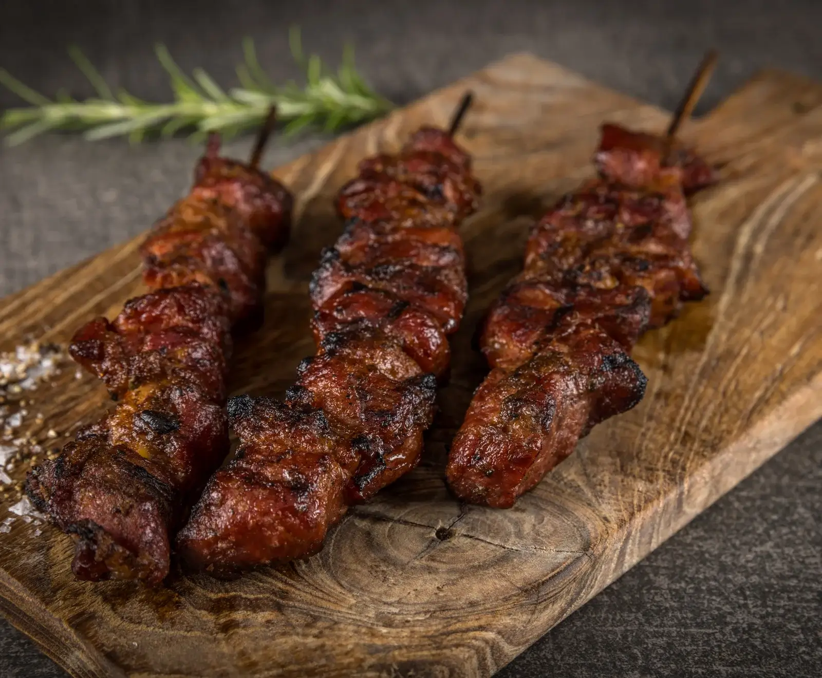 Mehrere Spieße mit gegrilltem, mariniertem Fleisch auf einem Holzbrett mit Rosmarin garniert
