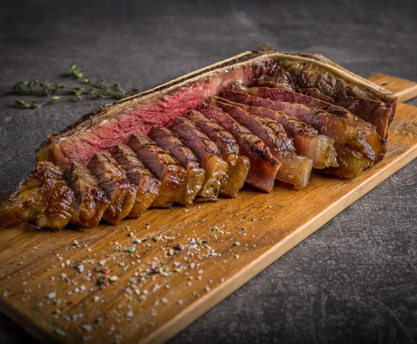 Ein aufgeschnittenes, medium-rare gegartes T-Bone-Steak auf einem Holzschneidebrett, bestreut mit grobem Salz und Pfeffer. Im Hintergrund sind frische Kräuter zu sehen.