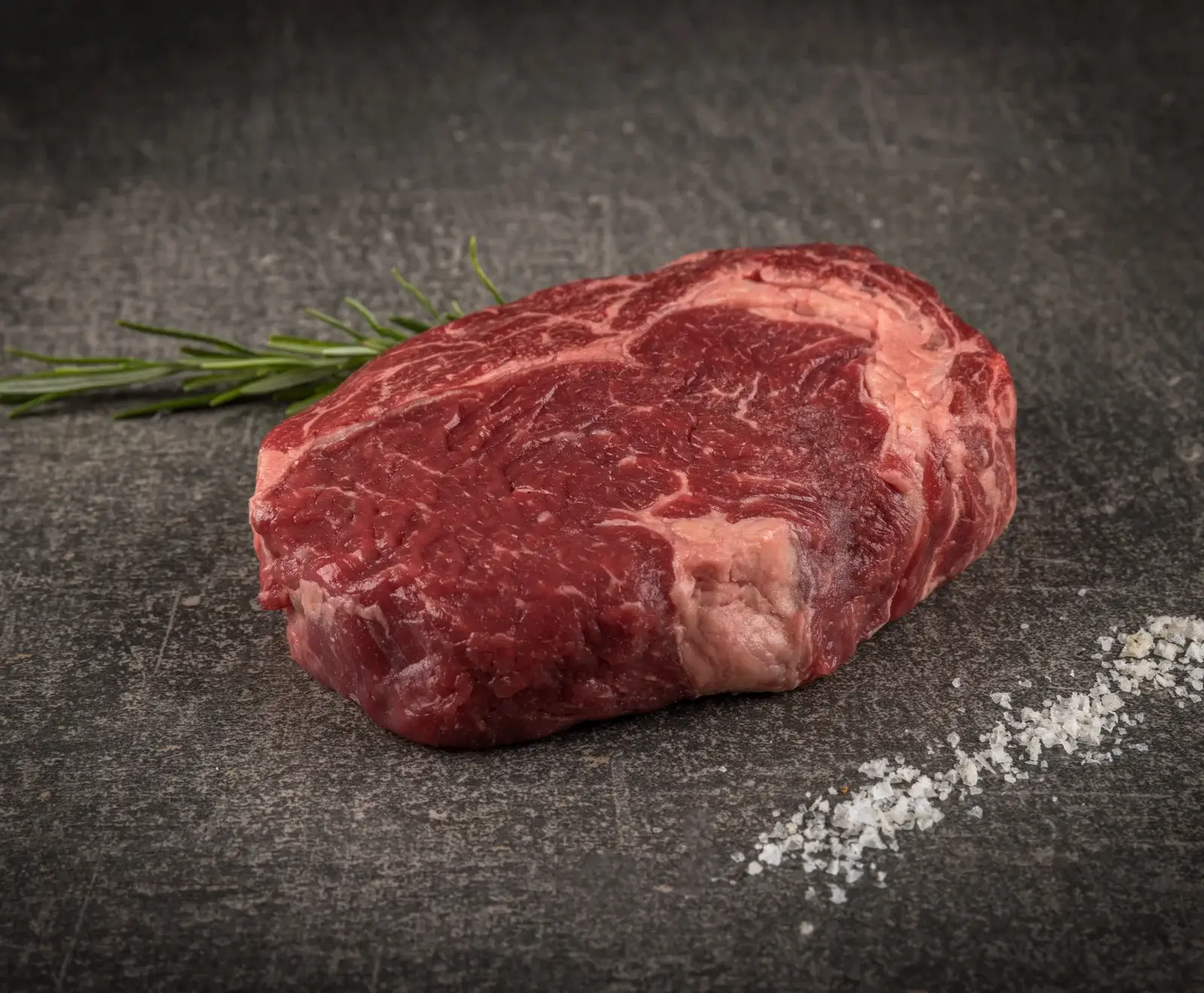 Ein rohes, marmoriertes Rindersteak auf dunklem Untergrund, mit einem Rosmarinzweig im Hintergrund und grobem Salz im Vordergrund.