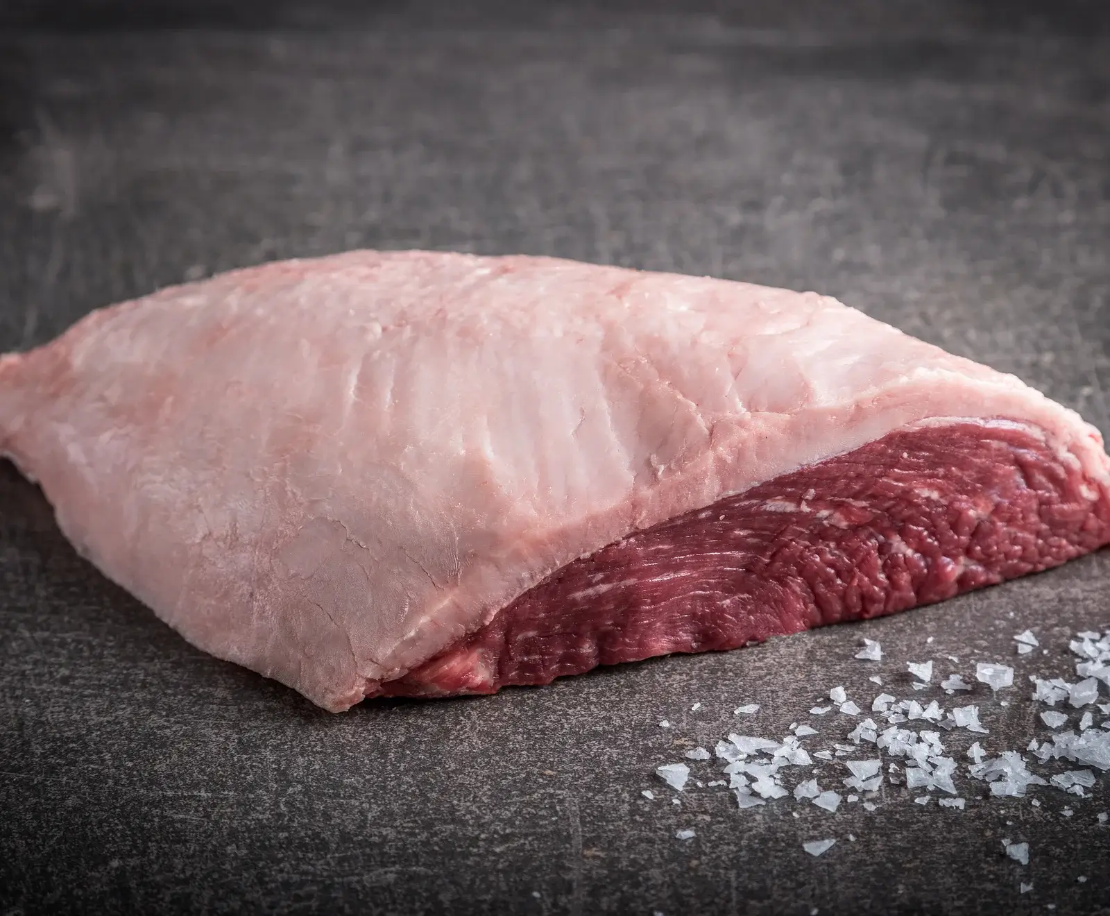 Rohes Schweinefleisch mit Salz auf dunklem Hintergrund, Nahaufnahme eines Fleischstücks mit Fettrand.