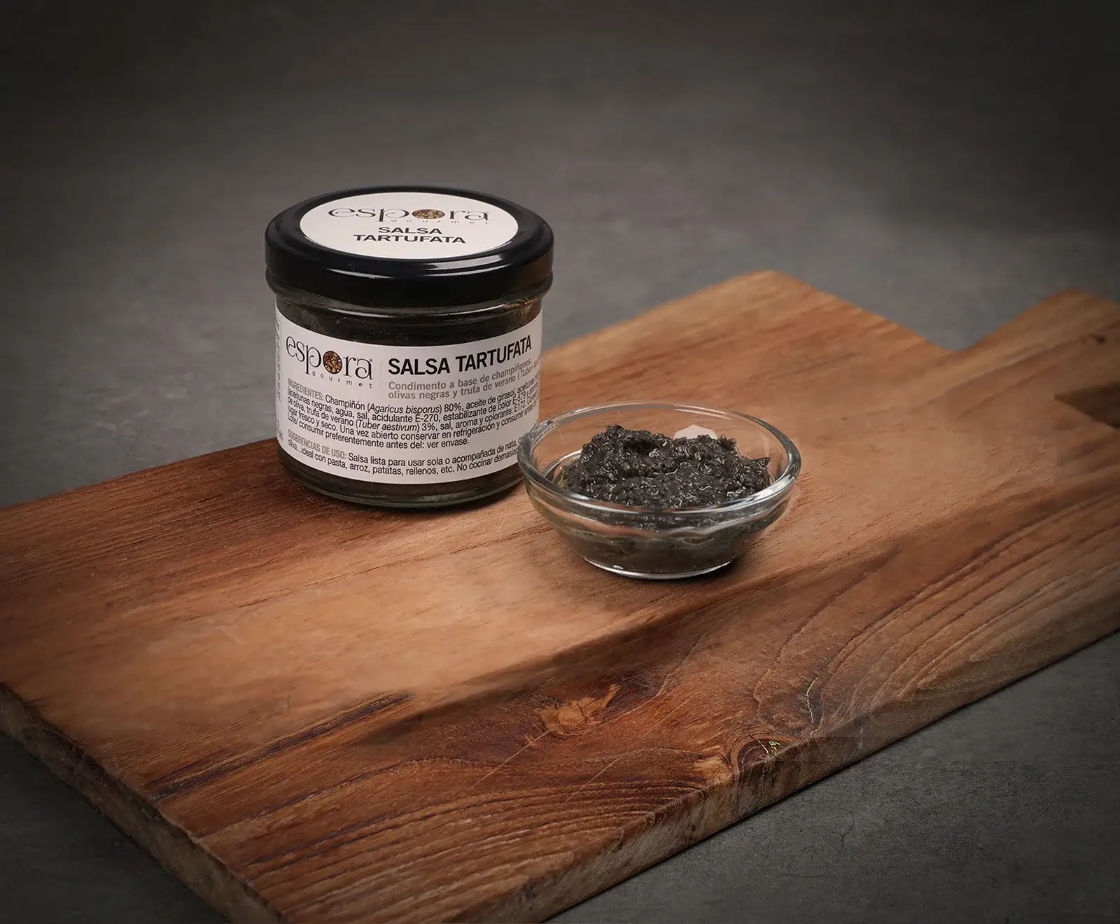 Ein Glas "espora gourmet Salsa Tartufata" und eine kleine Glasschale mit dunkler, grobkörniger Trüffelsauce stehen auf einem rustikalen Holzbrett vor einem dunklen Hintergrund.