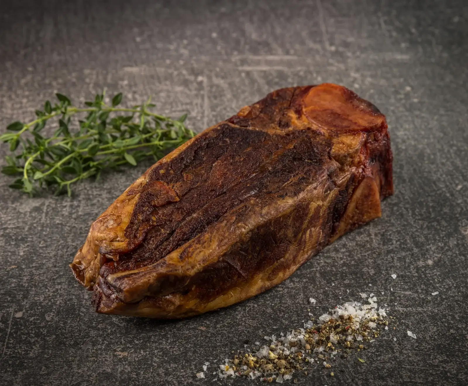 Ein großes, dunkelbraun gebratenes oder geräuchertes Fleischstück mit Knochen auf dunkler Steinplatte, mit frischem Thymian und grobem Salz und Pfeffer.