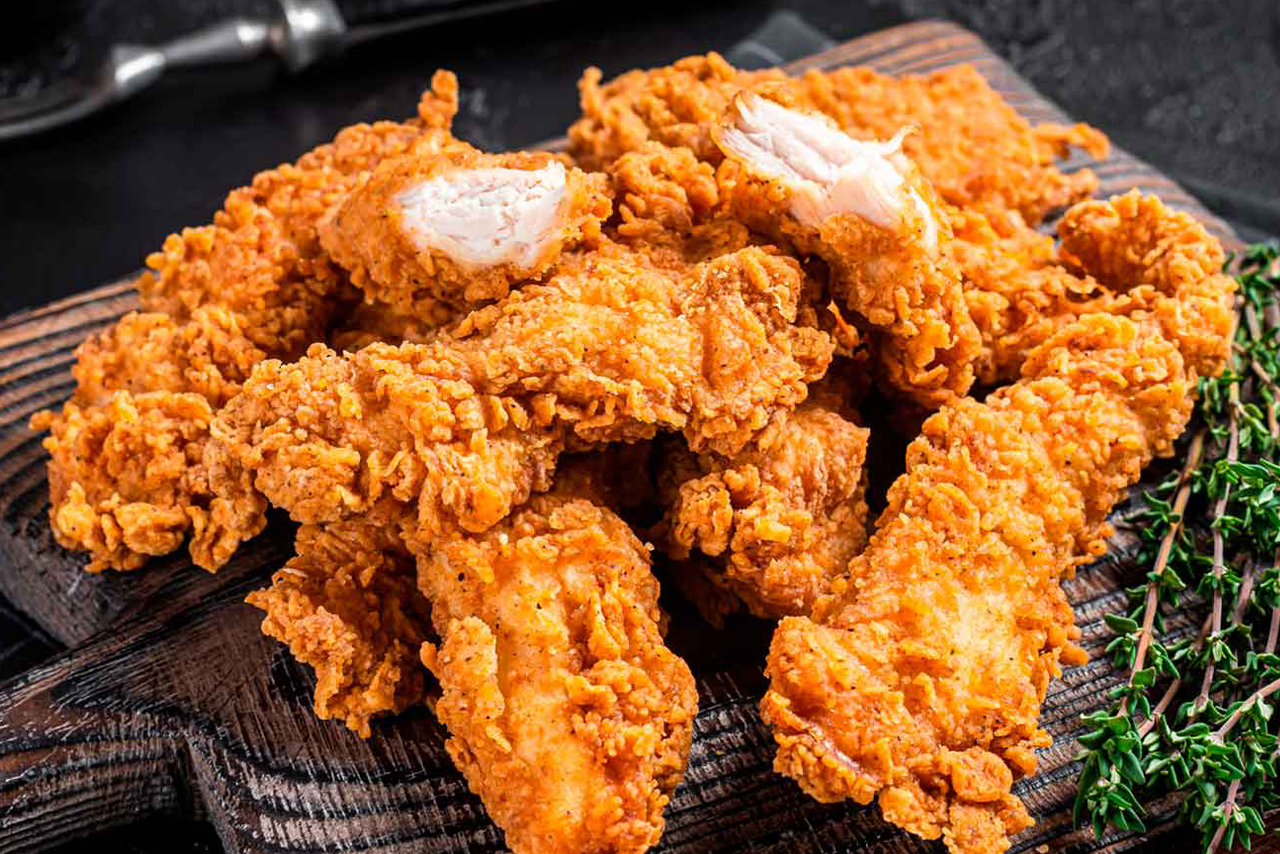 Ein Haufen goldbrauner, knuspriger Hähnchenstreifen auf einem dunklen Holzbrett, einige sind aufgebrochen und zeigen das weiße Fleisch. Garniert mit frischen Thymianzweigen.