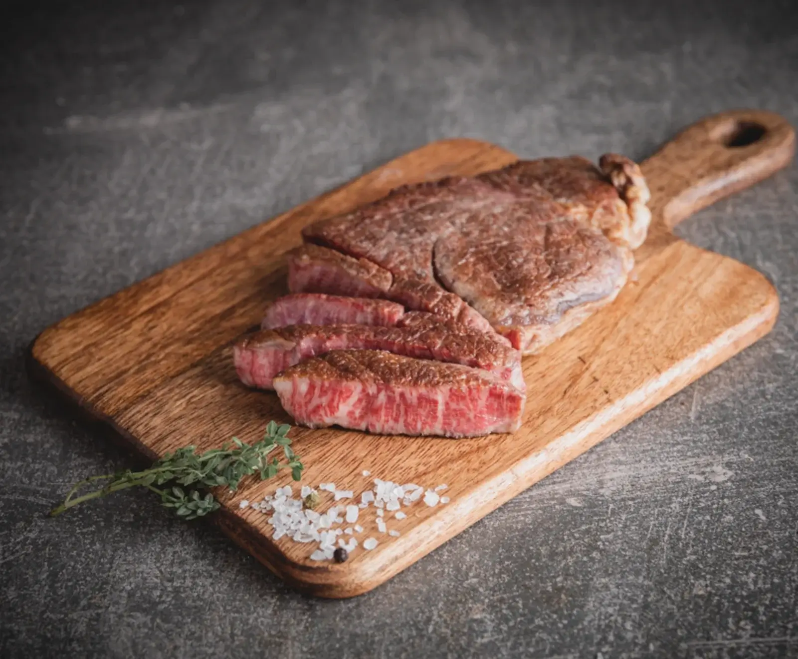 Ein saftiges, medium gebratenes Steak, teilweise in Scheiben geschnitten, liegt auf einem Holzschneidebrett. Daneben sind grobes Meersalz und ein Thymianzweig zu sehen. Der Hintergrund ist dunkelgrau.