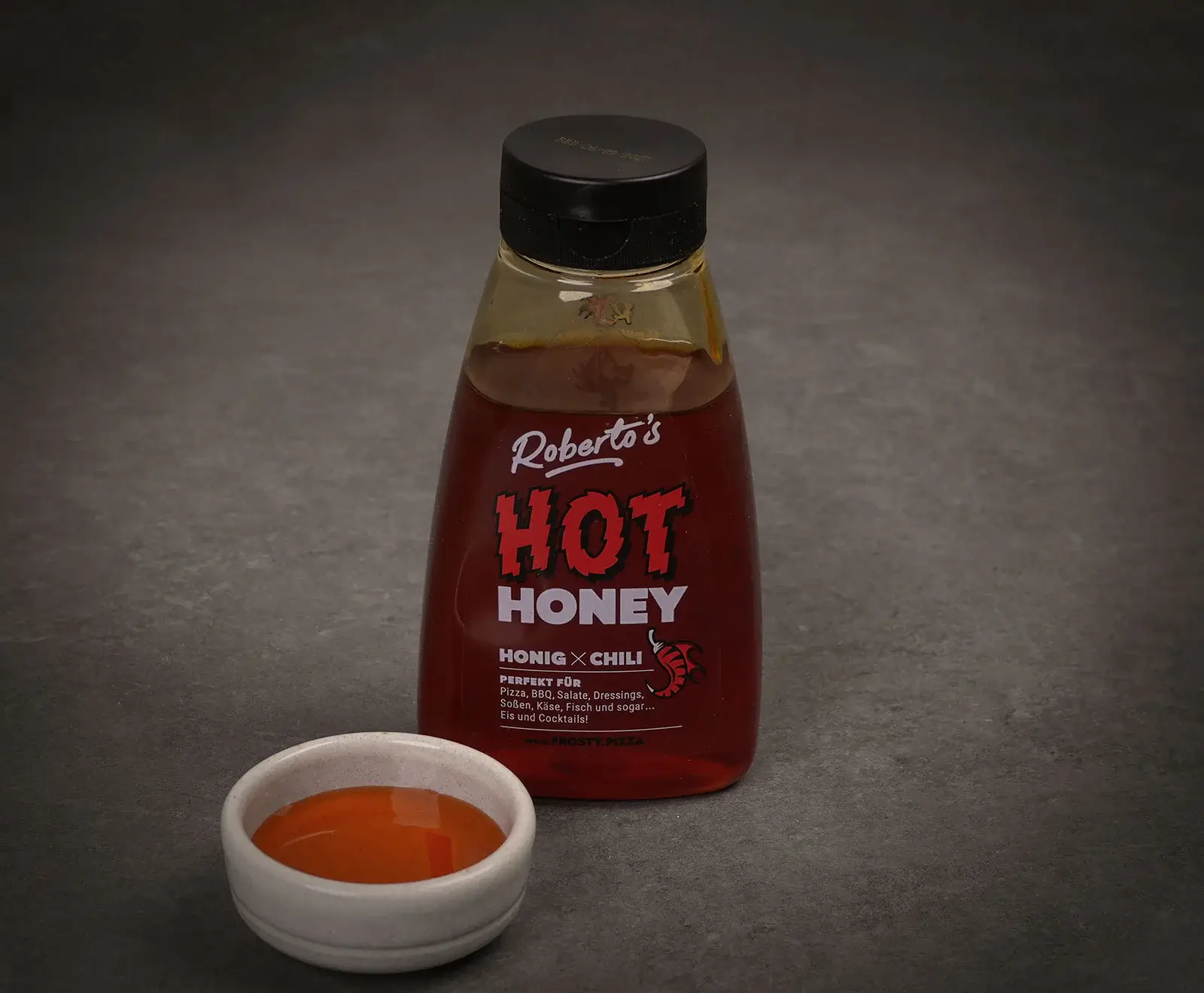 Eine Flasche Roberto's Hot Honey (Honig x Chili) mit rotem Inhalt und schwarzem Deckel steht auf einer dunklen Oberfläche. Daneben ist eine kleine weiße Schale mit etwas Hot Honey gefüllt.