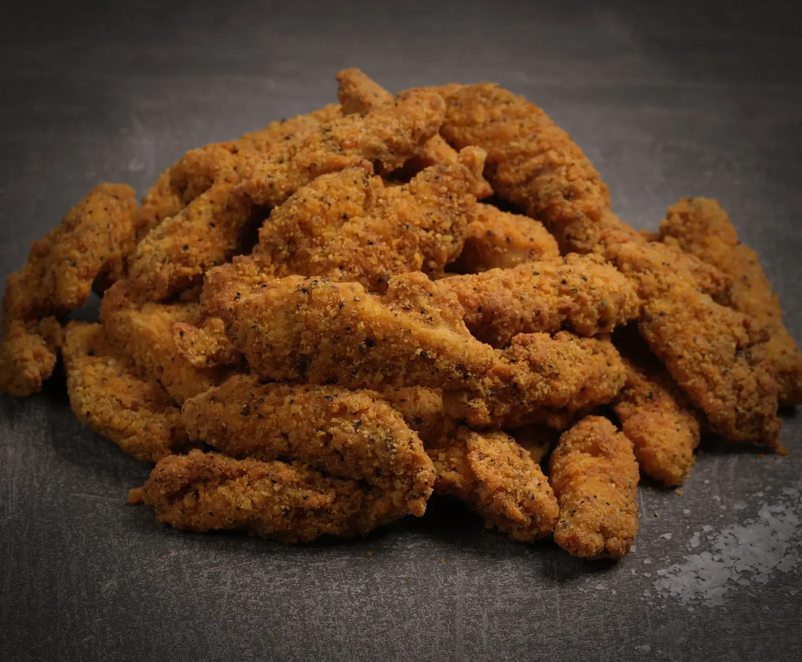 Ein Haufen knuspriger, goldbrauner Hähnchenstreifen, paniert und mit schwarzen Pfeffersprenkeln gewürzt, liegt auf einem dunklen, strukturierten Untergrund.