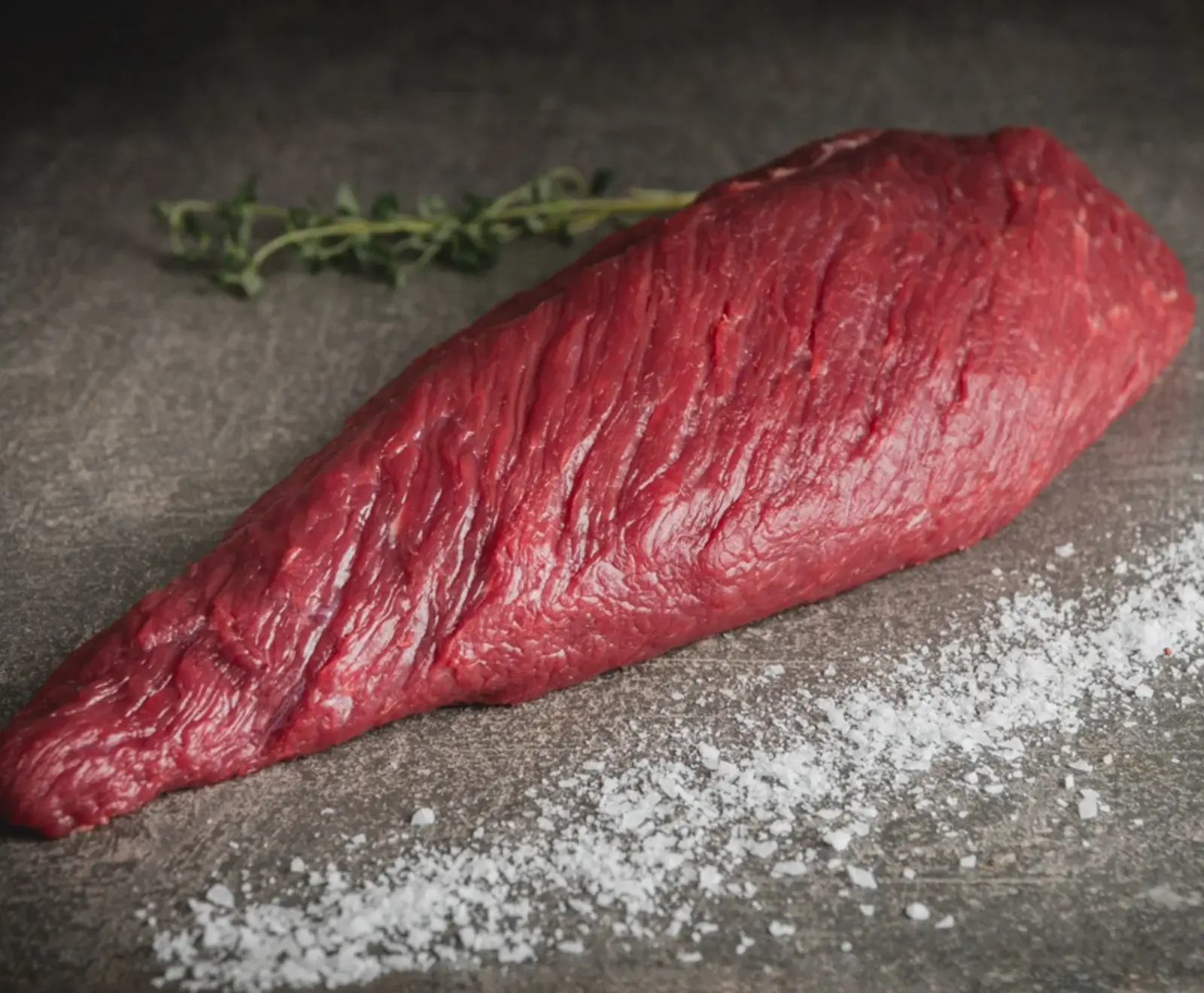 Ein rohes, dunkelrotes Stück Rindfleisch liegt auf einer dunklen, strukturierten Oberfläche. Grobes Salz ist im Vordergrund verstreut, und ein Thymianzweig ist im Hintergrund unscharf zu sehen.