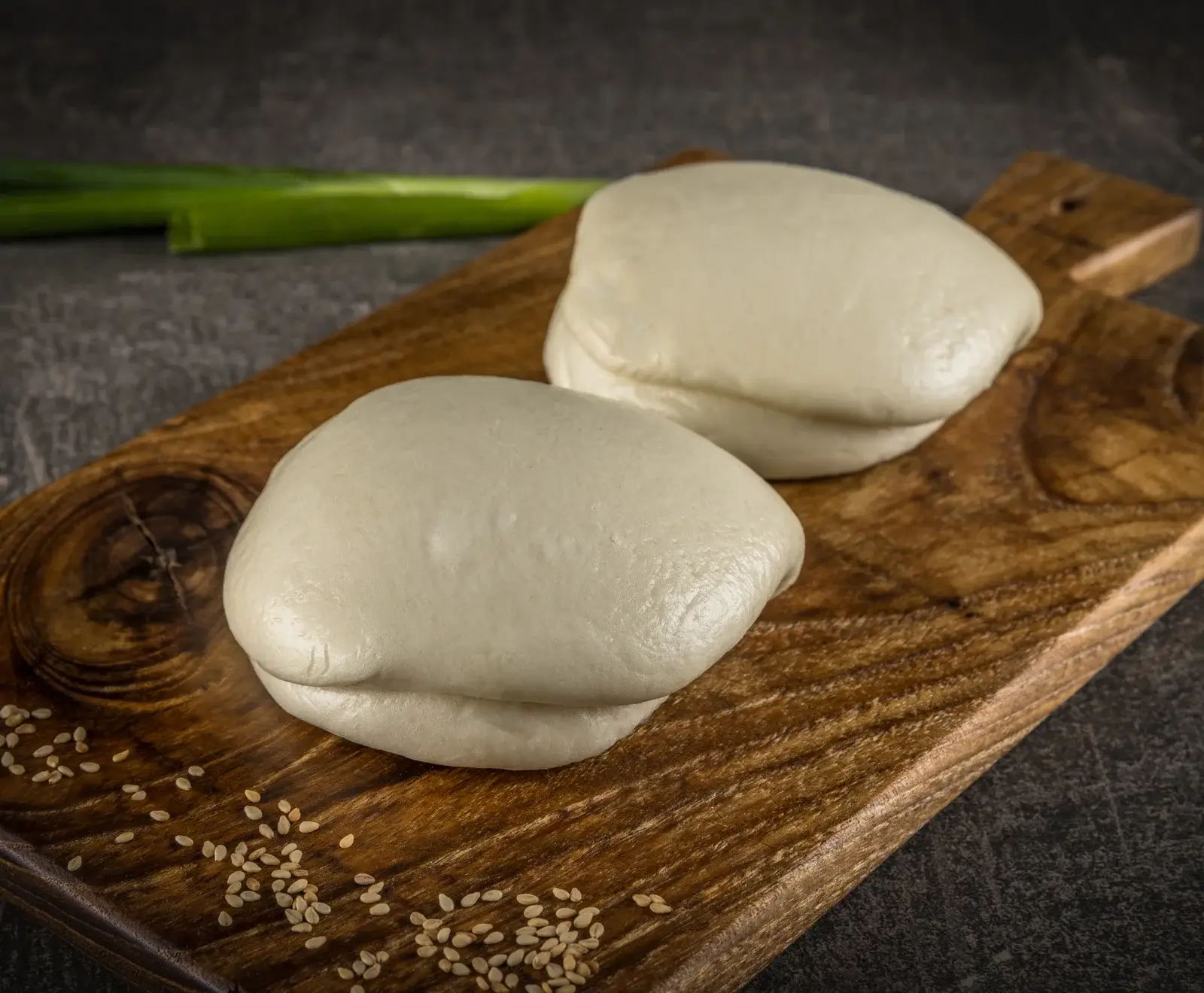 Zwei helle, gedämpfte Bao Buns auf einem Holzbrett mit Sesamsamen und Lauchzwiebeln im Hintergrund.