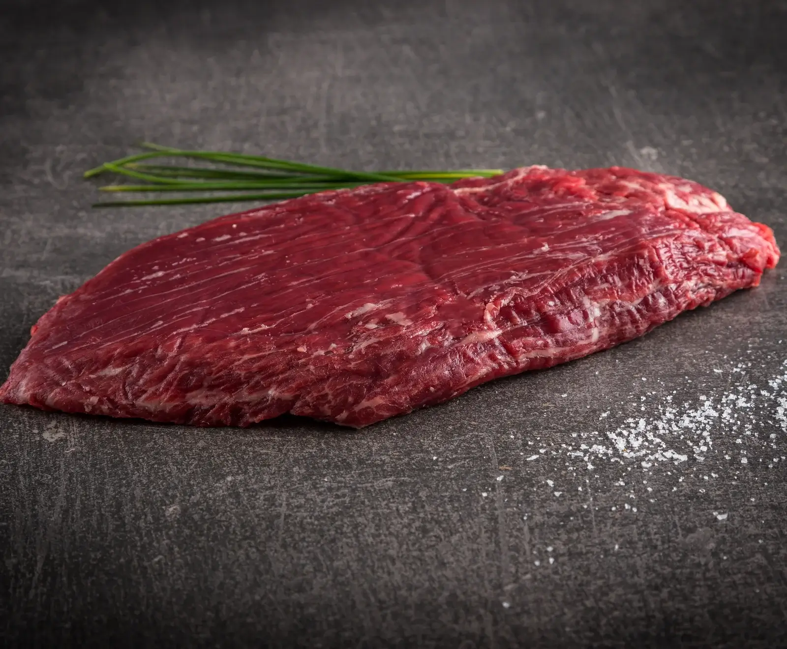 Ein rohes Flanksteak mit feinen Salzkristallen bestreut auf einer dunklen Oberfläche neben grünen Zwiebelstängeln.
