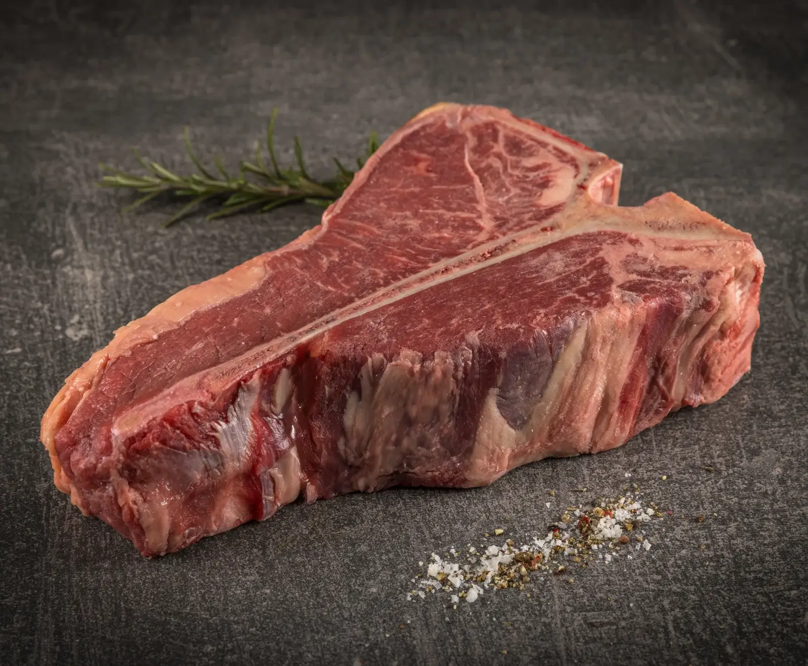 Ein rohes T-Bone-Steak liegt auf einer dunklen, strukturierten Oberfläche. Im Hintergrund ist ein Rosmarinzweig zu sehen, im Vordergrund eine Prise Salz und Pfeffer.