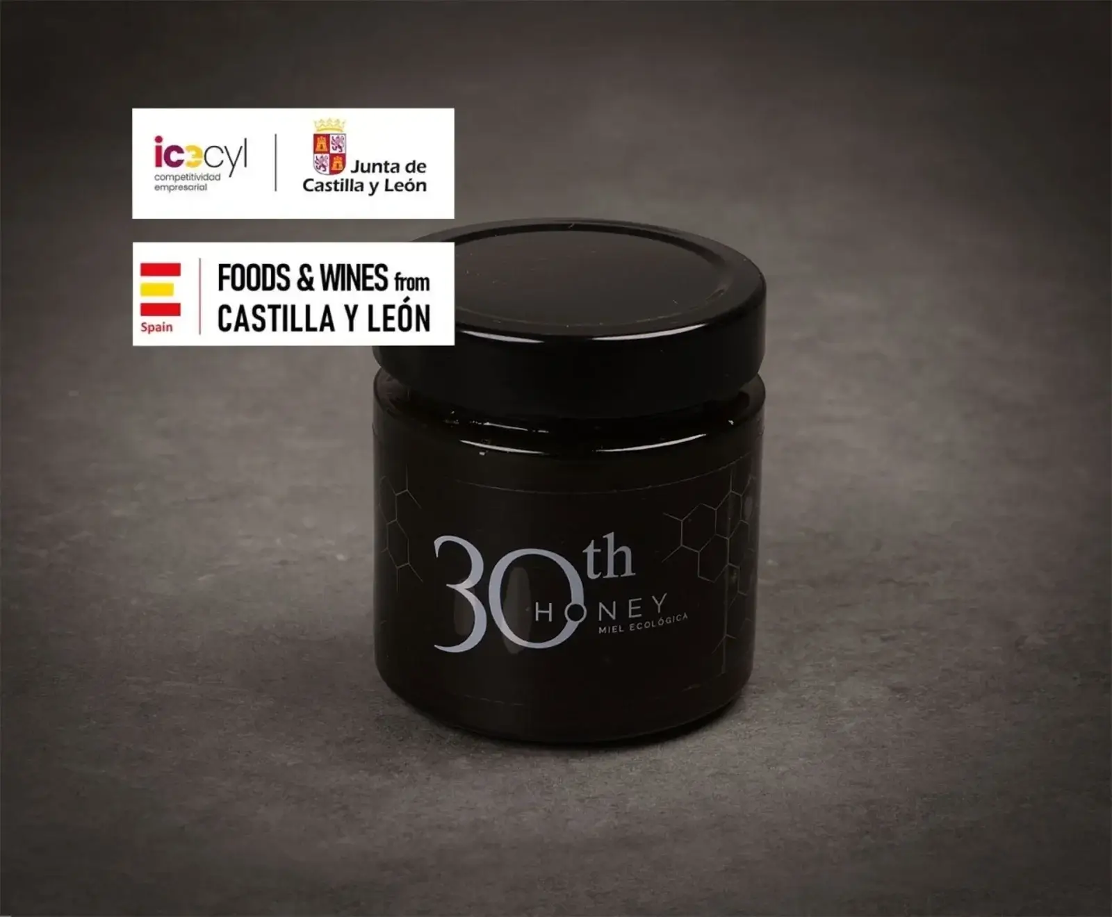 Ein dunkles Glas Honigglas mit schwarzem Deckel und dem weißen Aufdruck "30th HONEY MIEL ECOLÓGICA" auf einem dunklen Hintergrund. Oben links sind Logos von "Junta de Castilla y León" und "FOODS & WINES from CASTILLA Y LEÓN" zu sehen.