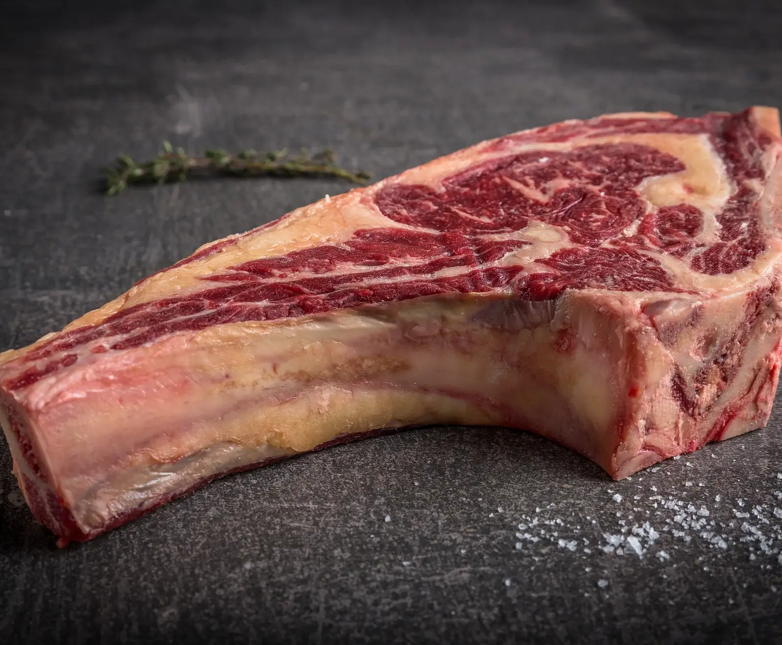 Rohes Ribeye-Steak mit Knochen, deutlicher Marmorierung und Fettrand, auf dunklem Untergrund mit Thymianzweig und grobem Salz. Bereit zur Zubereitung.