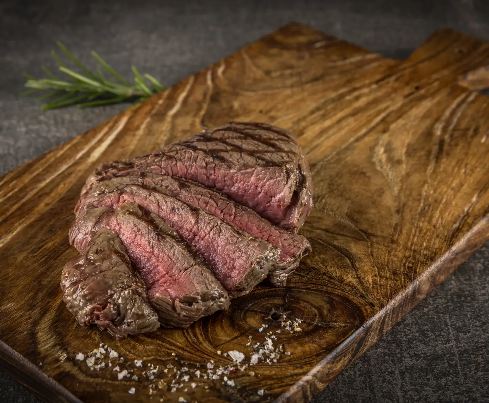 Ein aufgeschnittenes, medium-rare gegrilltes Steak mit rosa Kern und Grillstreifen liegt auf einem rustikalen Holzbrett, bestreut mit grobem Salz und Pfeffer. Ein Rosmarinzweig ist im Hintergrund.