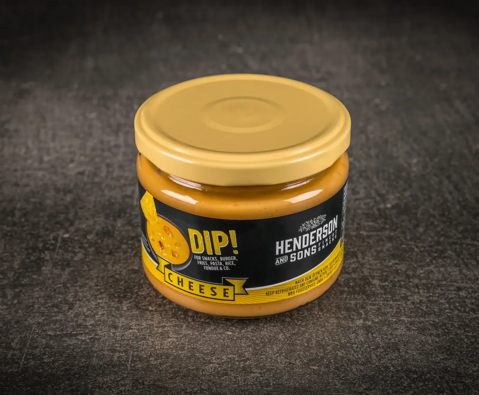 Ein Glas Henderson and Sons Käse-Dip mit gelbem Deckel und schwarzem Etikett, gefüllt mit orangefarbenem Dip, steht auf dunklem, strukturiertem Untergrund.
