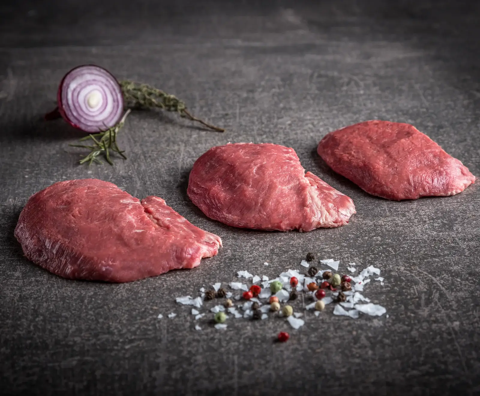 Drei rohe, rote Fleischstücke auf dunklem Stein, umgeben von einer halben roten Zwiebel, Rosmarin, Thymian, grobem Meersalz und bunten Pfefferkörnern. Bereit zum Kochen.