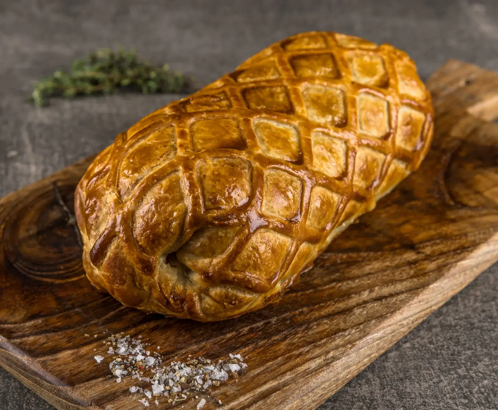 Ein goldbraunes, knuspriges Beef Wellington mit Rautenmuster auf einem rustikalen Holzbrett, garniert mit grobem Salz, Pfeffer und Thymian im unscharfen Hintergrund.