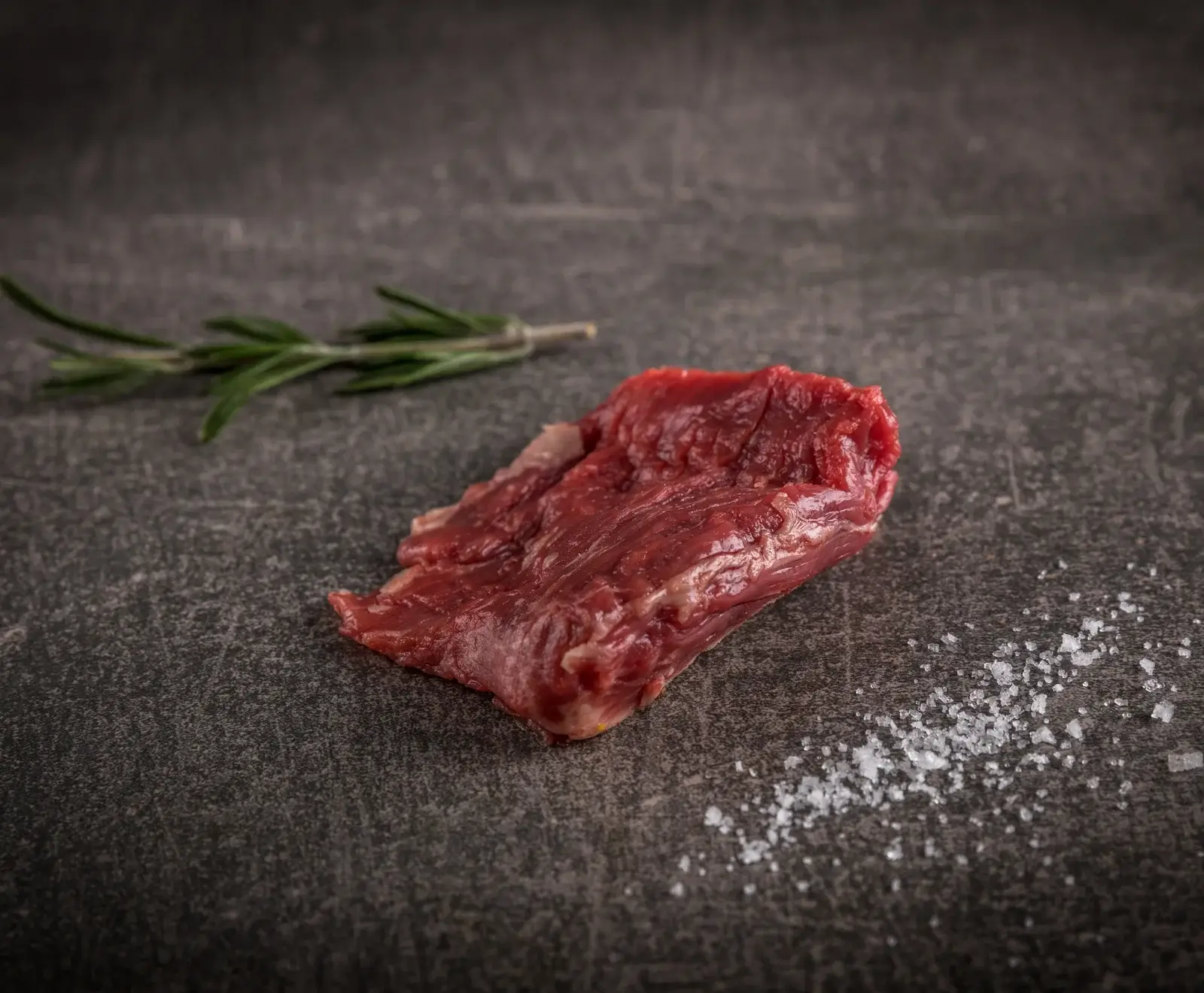 Ein Stück rohes, rotes Rindfleisch liegt auf einer dunklen Steinoberfläche. Links daneben ist ein Rosmarinzweig, rechts unten grobes Meersalz.