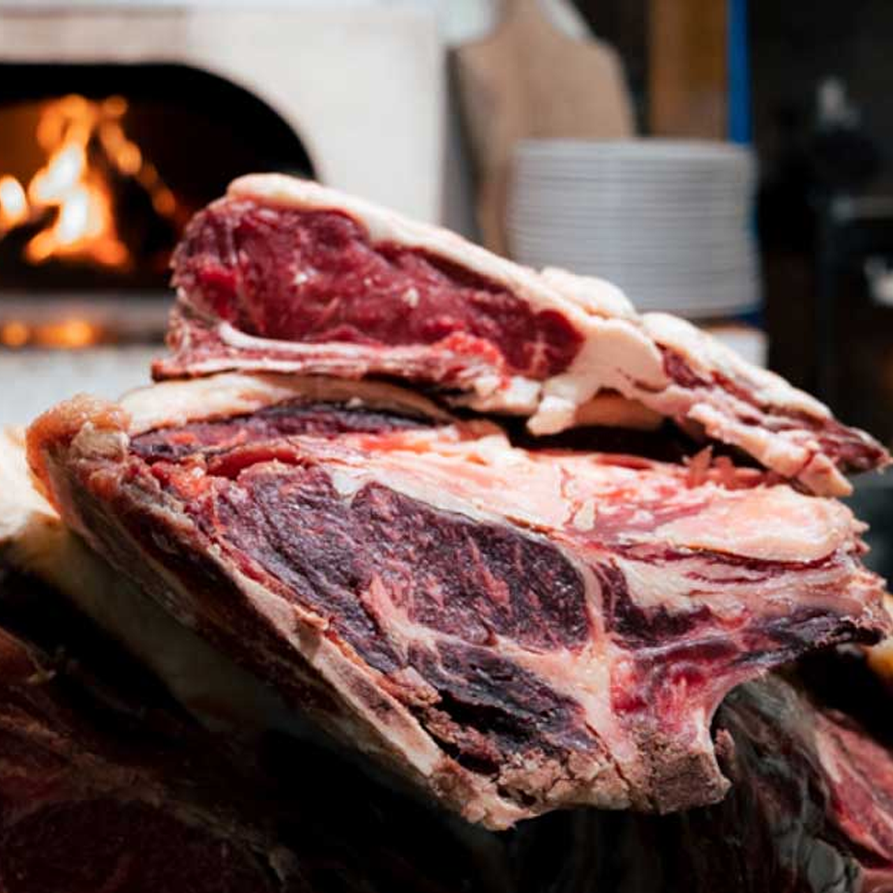 Nahaufnahme von mehreren großen Stücken rohem Rindfleisch mit Fett und Marmorierung, bereit zum Kochen. Im unscharfen Hintergrund ist ein Holzofen mit lodernden Flammen und ein Stapel Teller zu sehen.