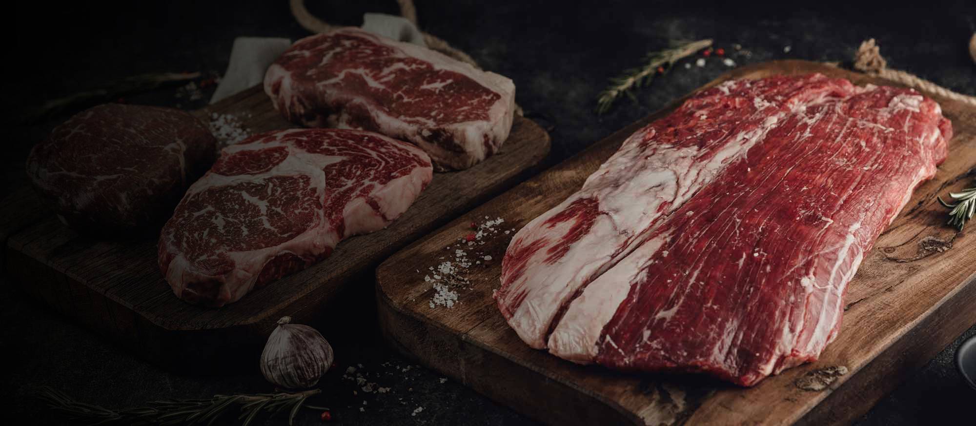 Rohes Rindfleisch: Zwei marmorierte Steaks und ein großes Stück Flanksteak auf Holzbrettern, umgeben von grobem Salz, Pfeffer, Rosmarin und Knoblauch auf dunklem Hintergrund.
