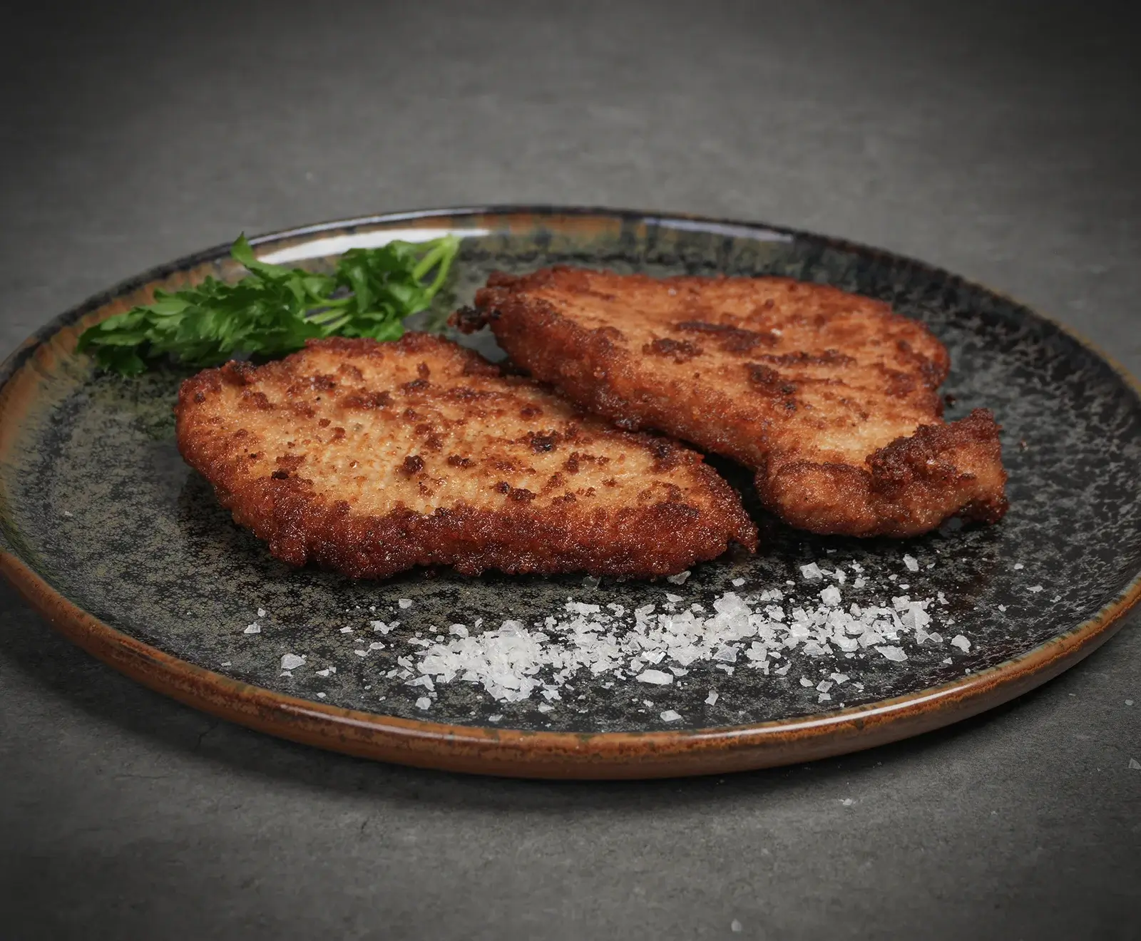 Zwei goldbraun panierte Schnitzel liegen auf einem dunklen, gesprenkelten Teller. Daneben sind frische Petersilie und grobe Meersalzflocken zu sehen. Der Hintergrund ist dunkelgrau.