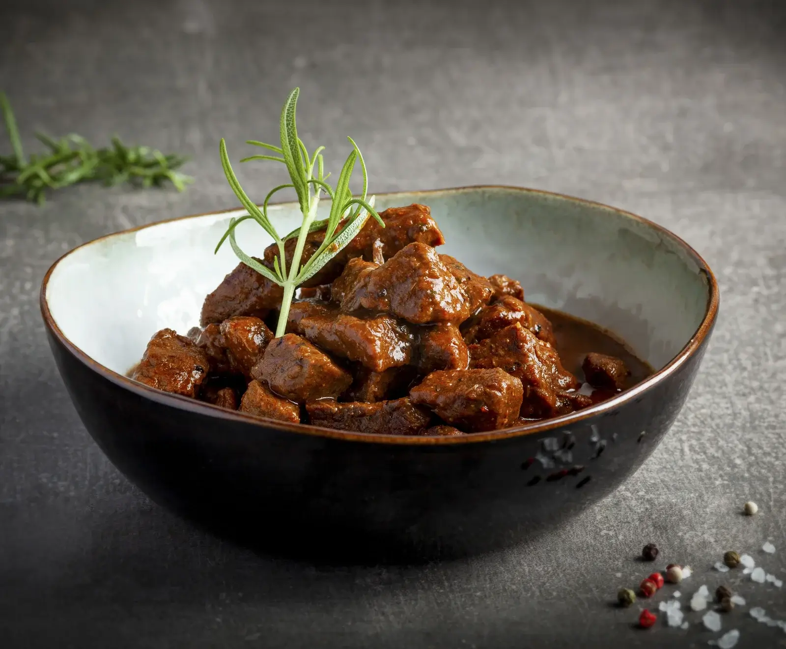 Nahaufnahme einer Schale mit herzhaftem Gulasch oder Fleischragout, verziert mit frischem Rosmarin. Im Vordergrund sind Pfefferkörner und grobes Salz zu sehen.