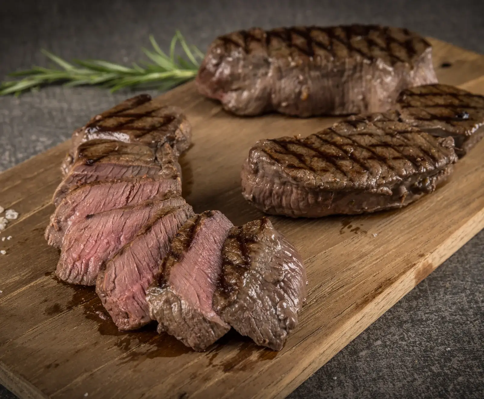 Nahaufnahme von drei saftigen, gegrillten Steaks auf einem Holzschneidebrett. Ein Steak ist in Scheiben geschnitten und zeigt eine medium-rare Garstufe. Rosmarin und grobes Salz sind im Hintergrund zu sehen.