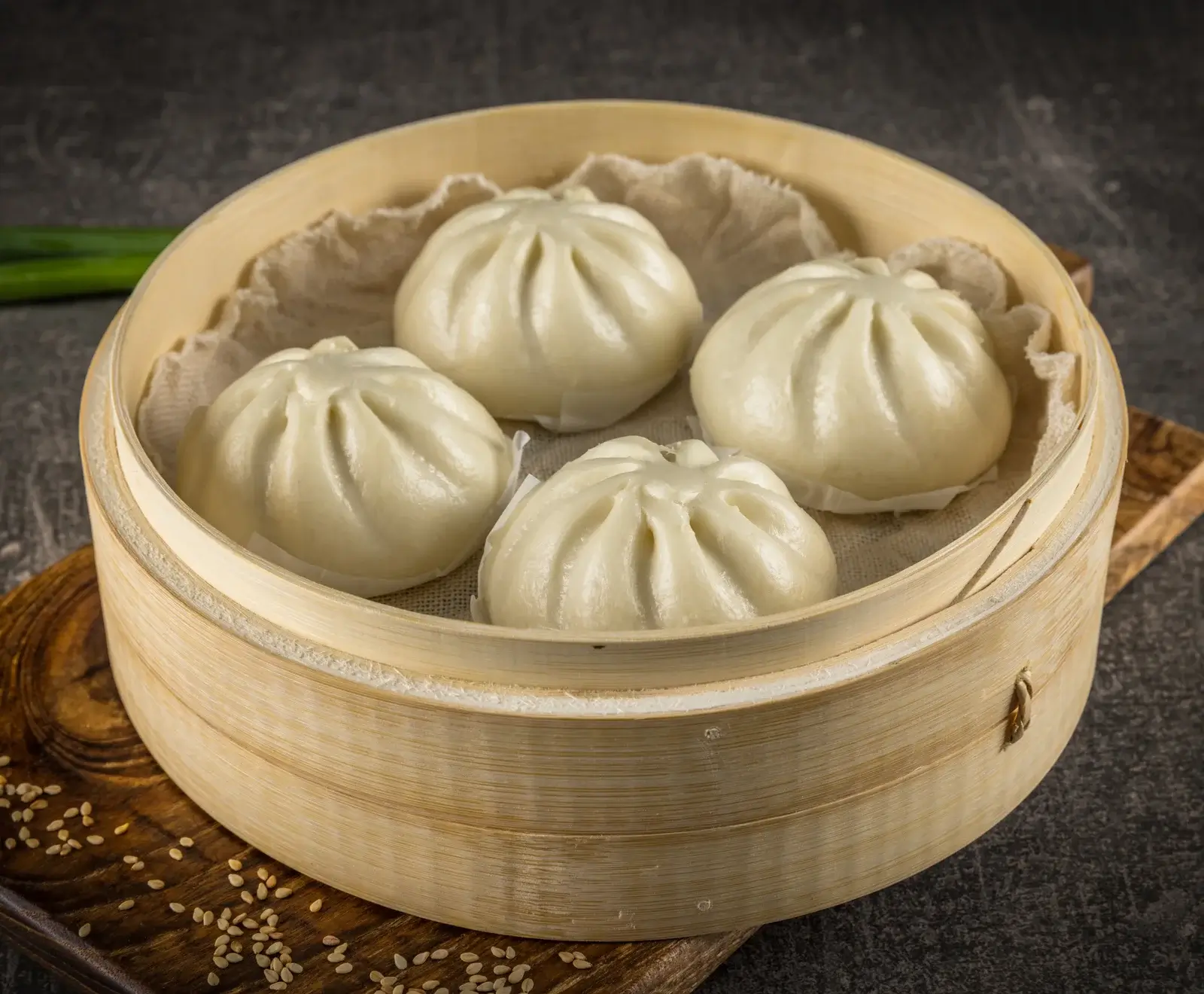 Vier gedämpfte, weiße Baozi-Brötchen in einem runden Bambusdämpfer auf einem dunklen Holzbrett mit Sesamsamen.