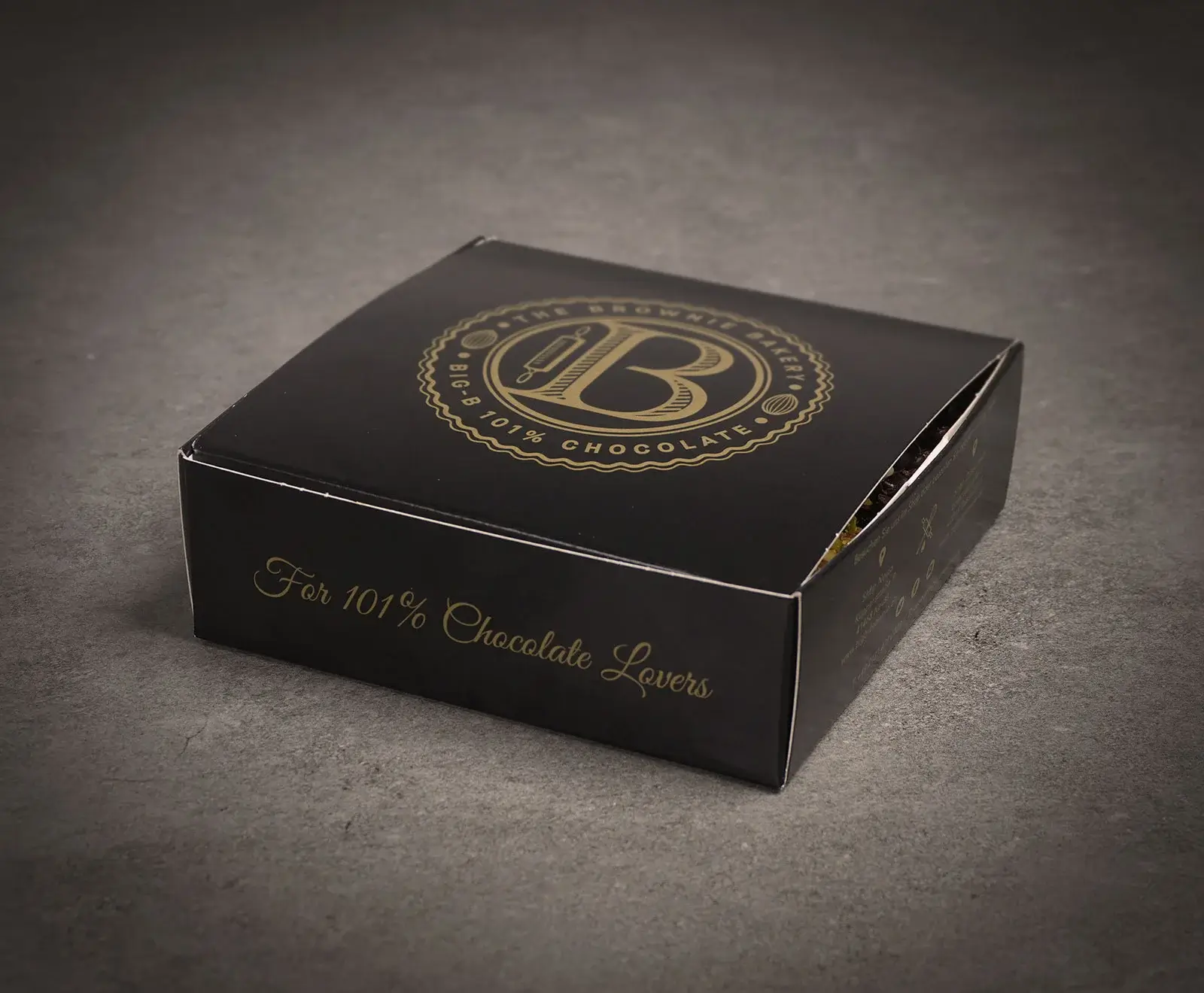 Schwarze Schachtel mit goldenem Logo von "THE BROWNIE BAKERY" und "BIG-B 101% CHOCOLATE" auf dem Deckel. Die Vorderseite trägt die Aufschrift "For 101% Chocolate Lovers". Die Box ist leicht geöffnet und steht auf einem dunklen Untergrund.