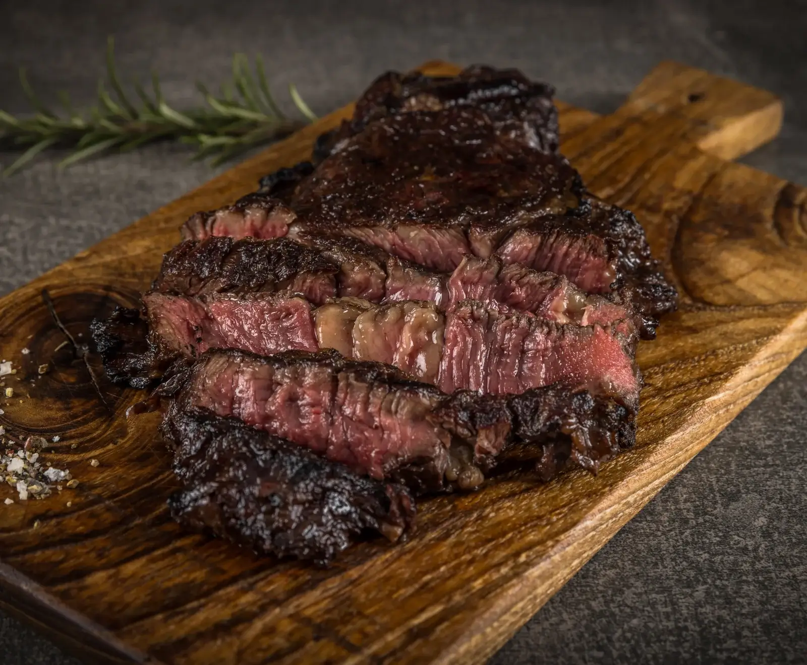 Ein perfekt gegartes, in Scheiben geschnittenes Steak mit rosa Kern und dunkler Kruste liegt auf einem Holzschneidebrett. Im Hintergrund sind Rosmarin und grobe Gewürze zu sehen.