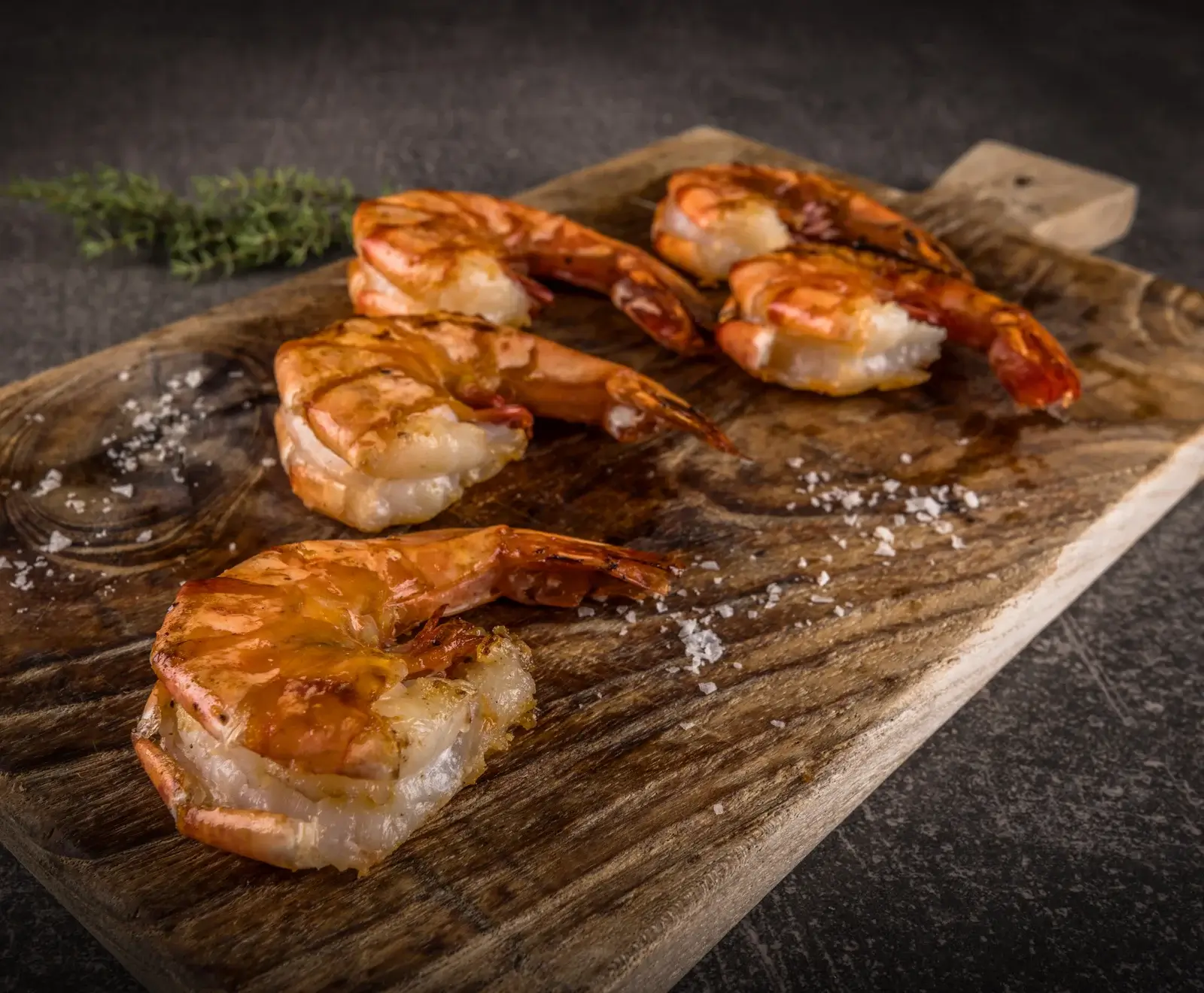 Fünf goldbraun gegrillte Garnelen mit Schwanz auf einem rustikalen Holzbrett, bestreut mit Meersalz. Im Hintergrund ein Thymianzweig auf dunklem Untergrund.