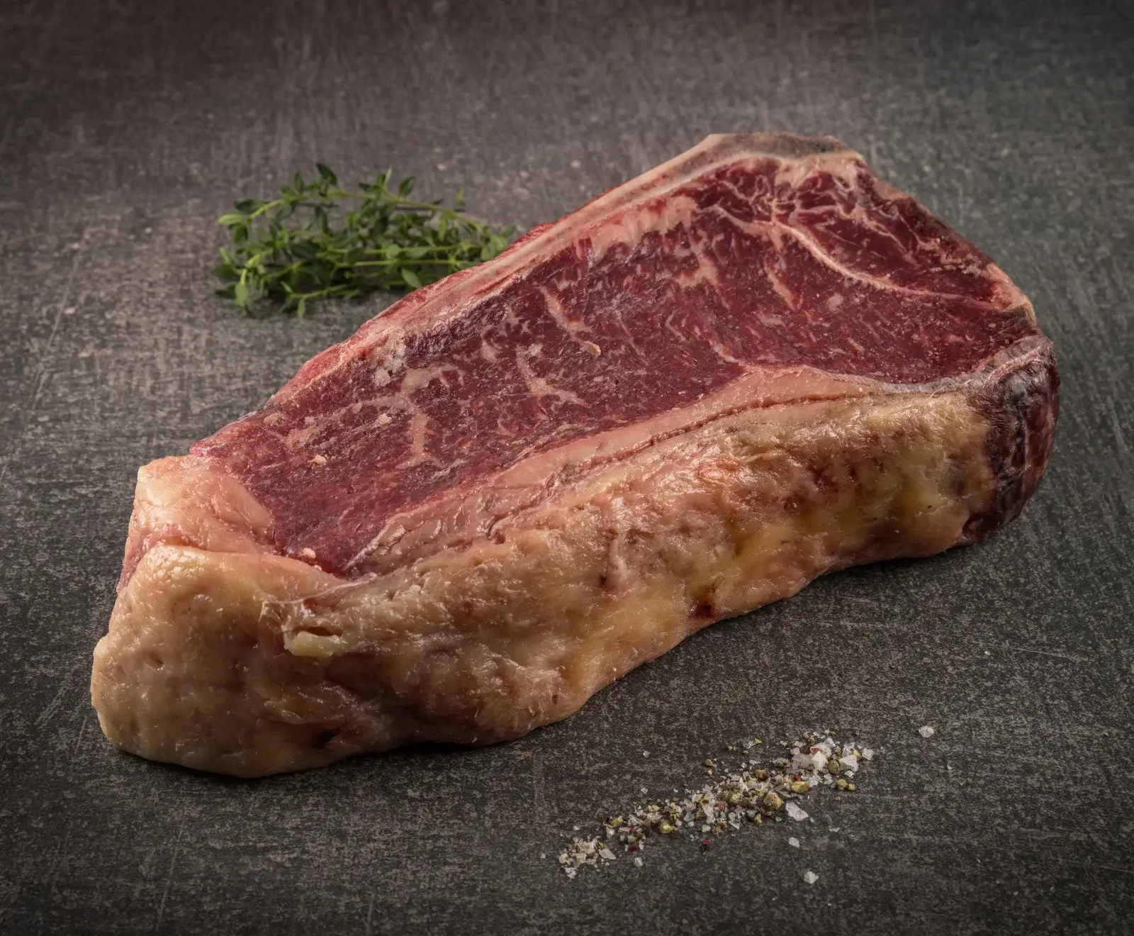 Ein rohes Rindersteak mit Knochen, starker Marmorierung und dickem Fettrand liegt auf dunklem Untergrund. Daneben sind frischer Thymian und eine Prise grobes Salz mit Pfeffer.